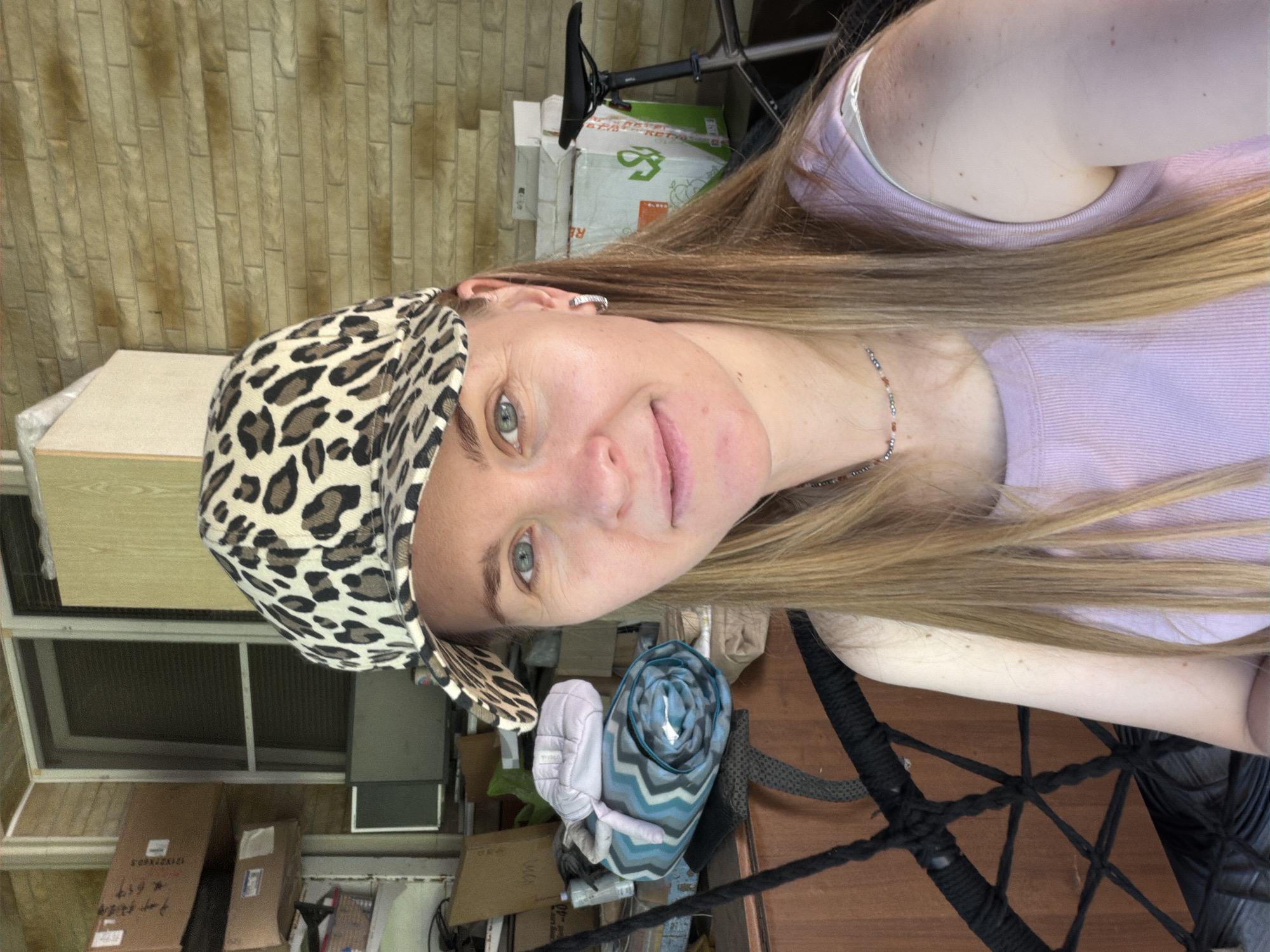 A woman is photographed from the chest up in a selfie. She is wearing a leopard print baseball cap, a light purple tank top, and a beaded necklace. Her hair is long and blonde. In the background, there are several cardboard boxes, a rolled-up blanket, and a bicycle with a black seat. The woman appears to be outdoors, with a brick wall visible behind her. The location is described as a storage area or porch, possibly in an urban setting.