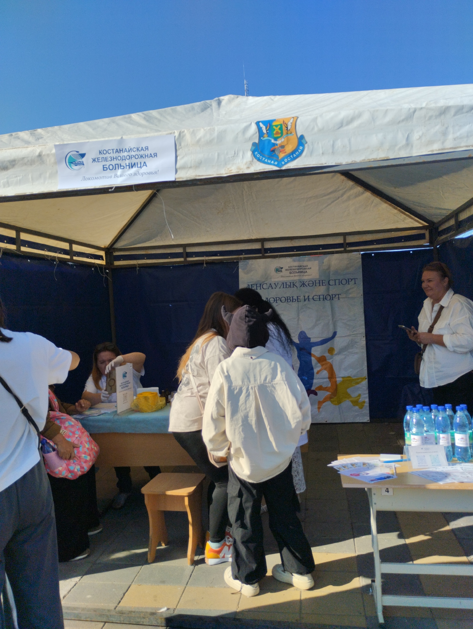 An outdoor event appears to be taking place under a white tent in Kostanay, Kazakhstan. A banner on the tent reads "KOCTAHAIIC KAR ЖЕЛЕЗНОДОРОЖНАЯ БОЛЬНИЦА" which translates to "Kostanay Railway Hospital" and a smaller tagline below says "Локомотив Вашего здоровья!" meaning "The locomotive of your health!". A coat of arms for Kostanay is also visible on the tent.
Inside the tent, a woman wearing gloves is seated behind a table, appearing to be assisting people. Several individuals, including a person wearing a white shirt and another in a white jacket and black pants, are standing near the table. A woman in a white shirt stands to the right of the tent, holding a phone. In front of the tent, a table is set up with bottles of water and some papers. The ground is paved with interlocking pavers. The sky is clear and blue.