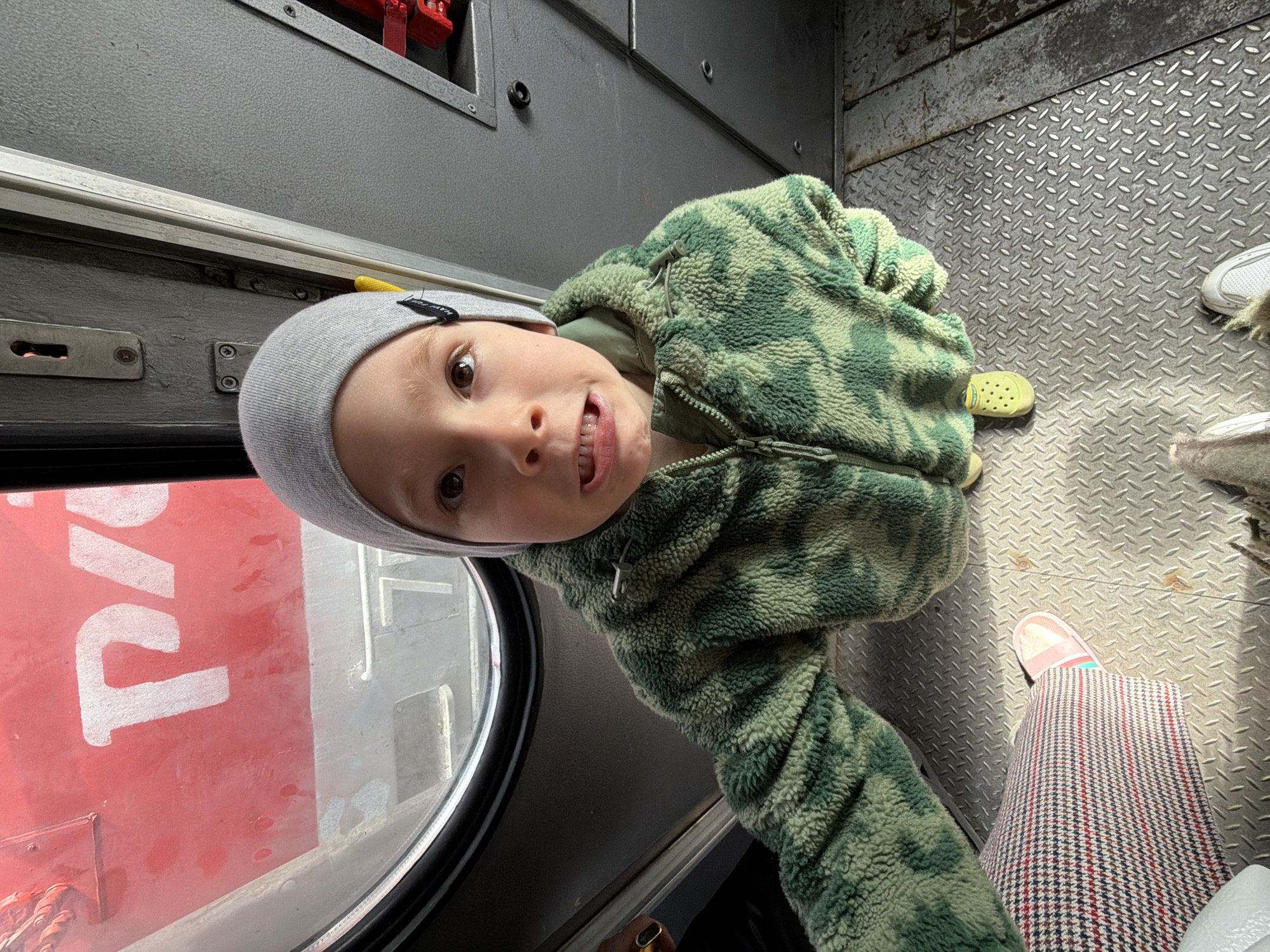 A young child, likely a boy, is the primary subject of the image, looking directly at the camera with a playful, open-mouthed expression. He is wearing a grey knit beanie and a green camouflage-patterned fleece jacket. His light brown eyes are wide, and a partial smile reveals his teeth.
The setting is the interior of what appears to be a utilitarian vehicle, possibly a train car or a maintenance compartment. The floor is made of diamond plate metal, and the walls are plain grey with visible bolts, a keyhole, and various fixtures. A round window on the left side of the frame offers a view of the outside.
Through the window, a large red surface with bold white Cyrillic text is visible. The letters "РУС" (RUS) are clearly distinguishable, which likely refers to "Russian" or "Russia," consistent with the location context of Liski, Russia, and commonly seen on Russian railway equipment.
In the lower right portion of the image, an adult's legs and feet are visible. The adult is wearing patterned trousers with red, blue, and white checks and light pink slip-on shoes. Next to them, a yellow Crocs-style shoe and another pair of white sneakers are on the metal floor.
The lighting suggests it is daytime. The child's expression conveys curiosity or amusement, captured in a candid moment within the industrial-looking interior of the vehicle.