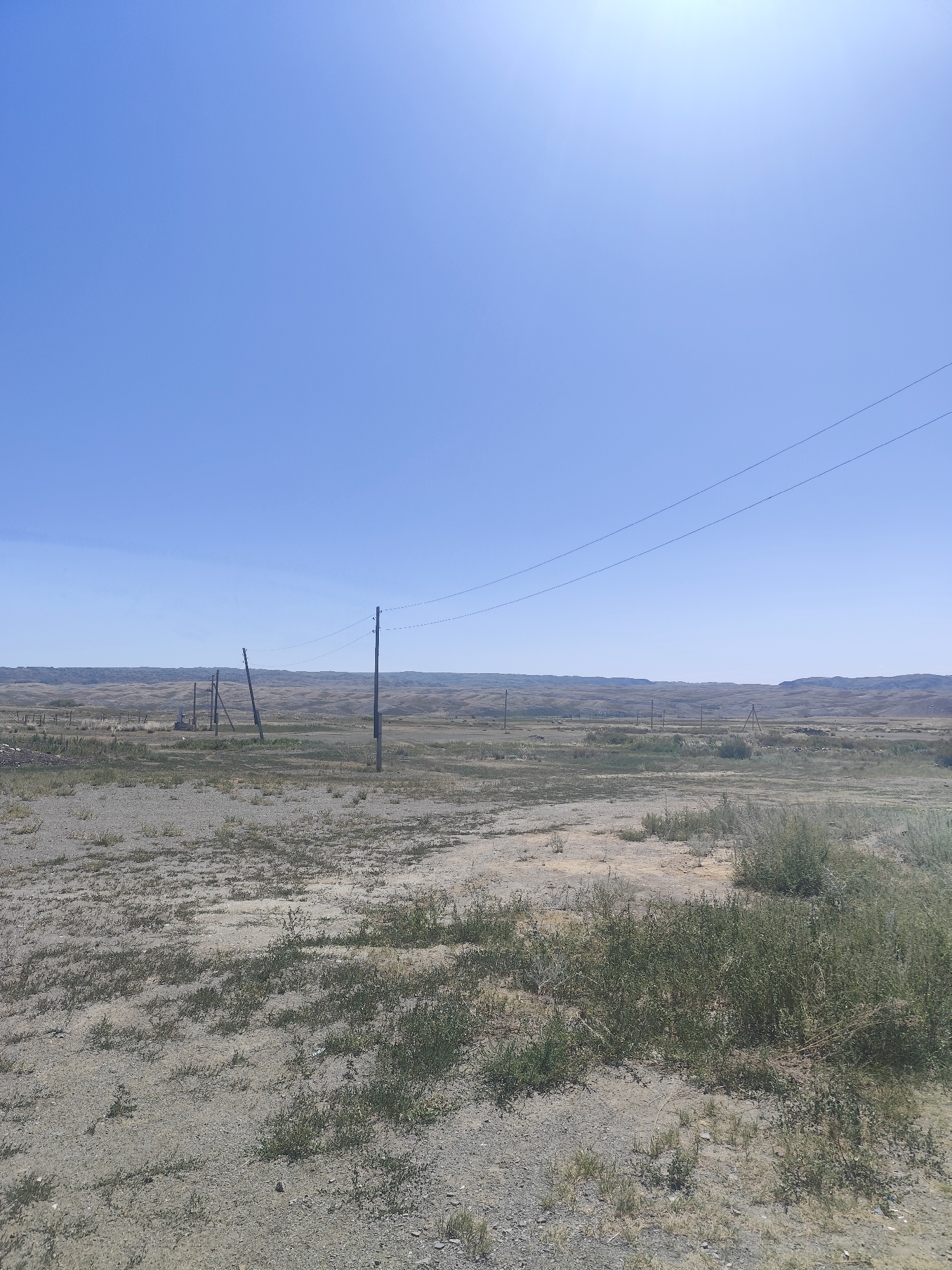The image depicts a vast, arid landscape under a clear blue sky, likely in the city of Aqbulaq Rural District, Kazakhstan. The terrain is mostly flat with gentle, undulating hills in the distance. The ground is covered in dry earth, gravel, and sparse, hardy vegetation, typical of a steppe or desert environment.

Several wooden utility poles with power lines stretched between them are visible, indicating the presence of infrastructure in this remote area. Some of the poles appear aged and weathered. In the mid-ground, there's a collection of these poles, some leaning precariously, suggesting either neglect or ongoing maintenance. A low, rustic fence can be seen on the left side of the frame, bordering a small, indistinct area.

The overall impression is one of a sparsely populated, open expanse, characterized by its natural, untouched beauty and the utilitarian presence of power lines. There are no people or animals visible in the image. The lighting suggests it is daytime, with the sun high in the sky, creating a bright, slightly washed-out effect towards the upper right corner.