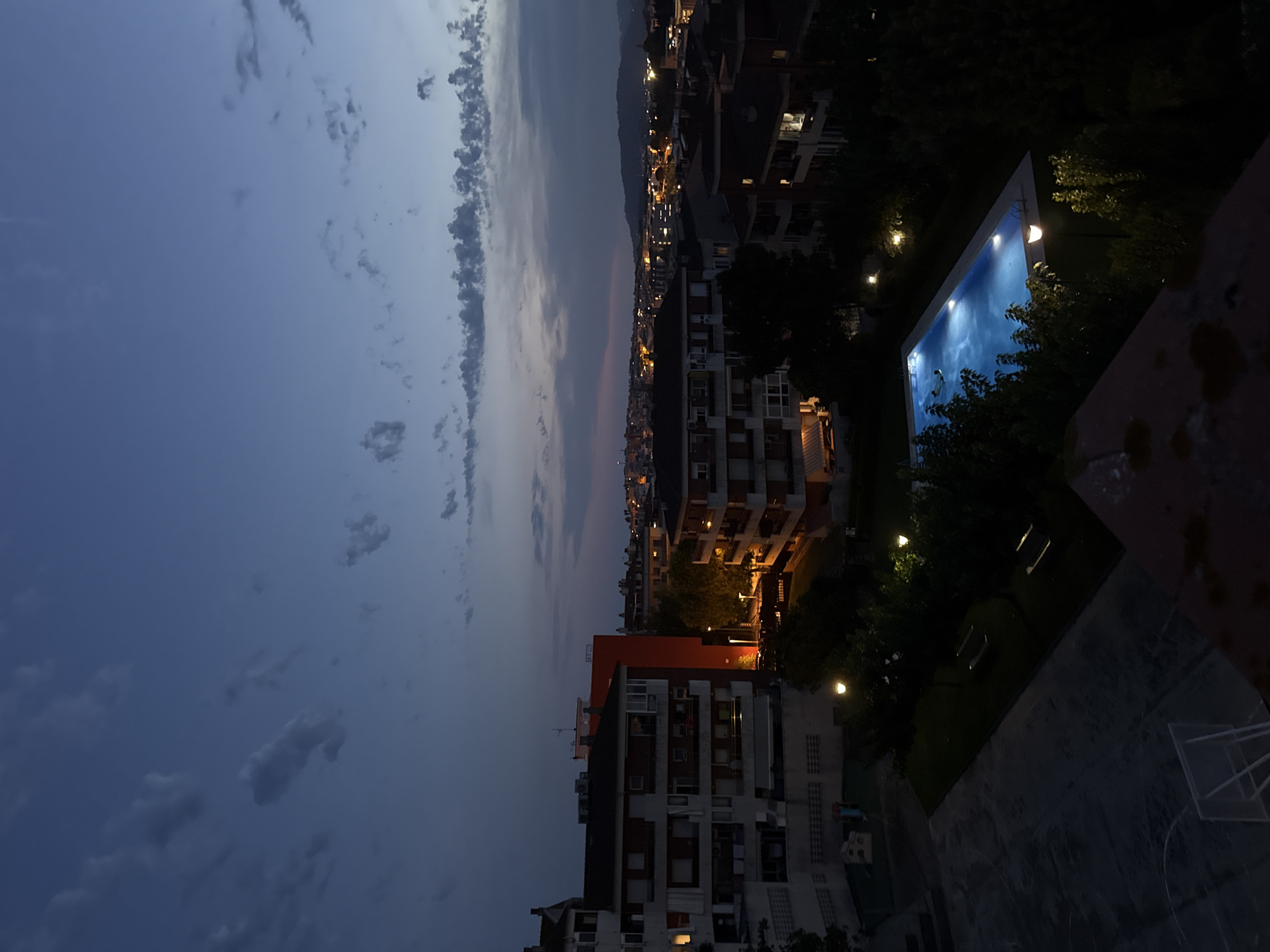 The image captures a twilight scene over a residential area in Blanes, Spain. In the foreground, a dark, partially visible rooftop area with some red brick detailing leads the eye towards the main subject of the image. A rectangular swimming pool, illuminated by internal lights, dominates the middle ground. The water within the pool is a vibrant blue, reflecting the light source. Two figures are visible in the pool, seemingly swimming or wading.
Surrounding the pool are lush green trees and manicured lawns. Several lampposts with glowing lights are strategically placed around the pool and in the adjacent park area, casting pools of light onto the grass and walkways. Beyond the immediate garden area, multi-story apartment buildings are visible, their windows dimly lit, suggesting evening hours. The buildings are a mix of white, beige, and red facades, typical of urban architecture.
In the distance, the city sprawls towards the horizon, with numerous lights twinkling from buildings and streetlights, indicating a settled urban environment. A range of hills or mountains forms the backdrop, silhouetted against the darkening sky. The sky itself is a gradient of deep blue to lighter hues near the horizon, with wispy clouds scattered across it, tinged with the last remnants of daylight or artificial light. The overall atmosphere is one of calm and serenity as day transitions into night.
