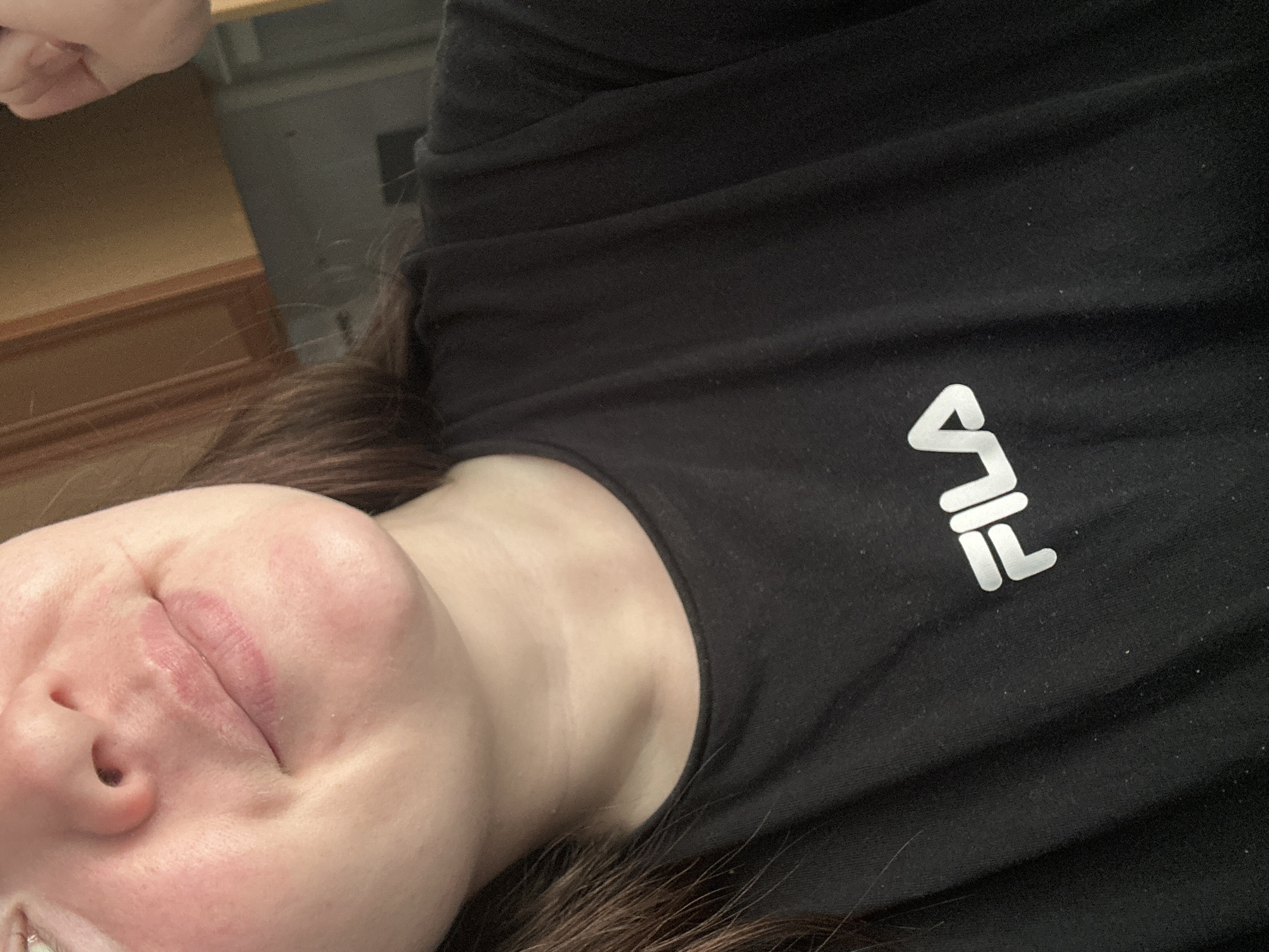 This is a close-up, over-the-shoulder shot of a person lying down. The subject is wearing a black t-shirt with the white Fila logo visible on the right side of the chest. The person's face is partially visible in the lower left of the frame, with their eyes closed and lips slightly parted. Their skin appears fair with a slight pinkish hue. Dark brown hair is visible around the subject's head and neck. The background consists of a wooden surface and a wall, suggesting an indoor setting. The lighting appears to be artificial, casting soft shadows. No specific activity is discernible other than resting. The scene is a personal, indoor environment, possibly a home, in Chaikovsky, Russia.
