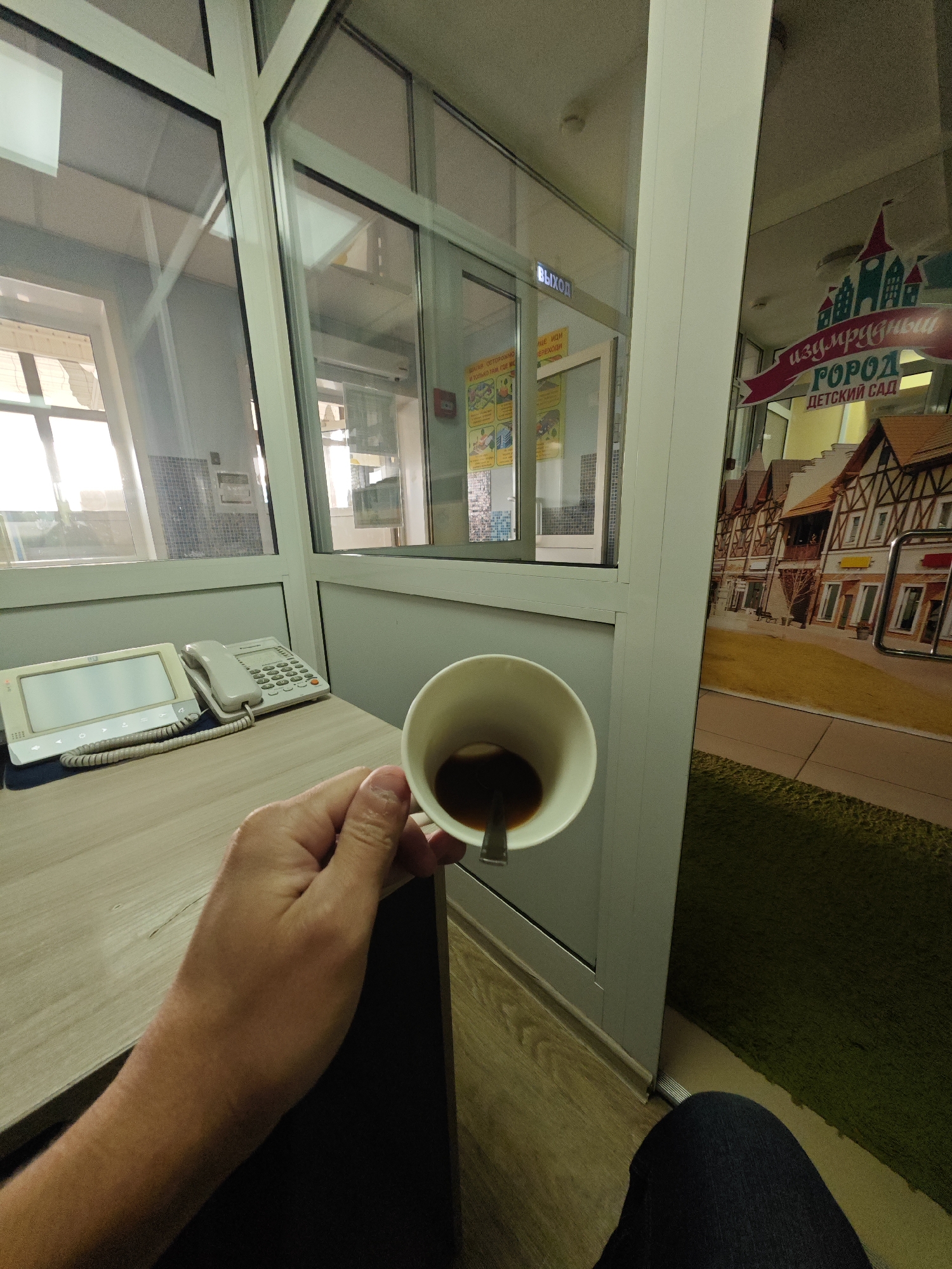 The image shows a first-person perspective of a hand holding a white mug filled with coffee. The mug has a spoon inside. The scene appears to be an office or reception area. In the background, there is a desk with a white telephone and a tablet device. Large windows reveal sunlight and reflections of the interior. On the right side of the image, a colorful mural depicting buildings and a banner with Russian text that reads "Изумрудный город детский сад" (Emerald City Kindergarten) is visible, suggesting the location is a kindergarten. The flooring is wooden and a green carpet. The context of Сухово, Russia, indicates this scene likely takes place within a building in that city.