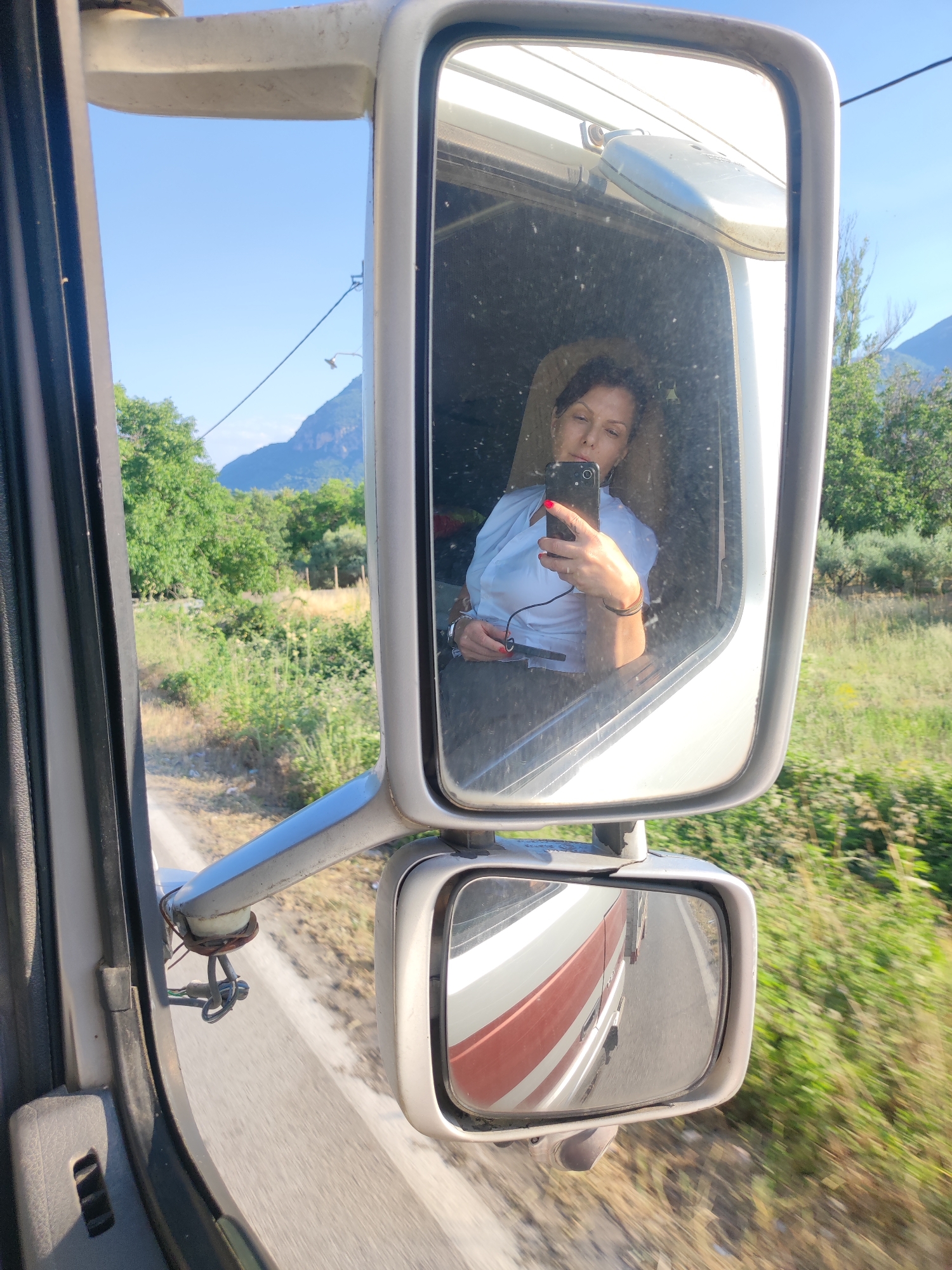 The image, taken in Kalabaka, Greece, captures a reflection of a woman in the side mirror of a truck. The woman is looking at her phone, which she holds in her right hand as she takes a selfie. She is wearing a light blue collared shirt and has dark hair. Her left arm is resting on her lap. The scene outside the truck's window, visible in the background of the mirror's reflection, shows a rural landscape with green trees, dry grass, and mountains in the distance under a clear blue sky. The sun appears to be shining, suggesting it is daytime. The smaller mirror below the main one reflects the side of the truck, showing a red and white stripe pattern. The overall impression is a moment of rest or a break from driving, with the woman capturing a personal moment against the backdrop of the Greek countryside.