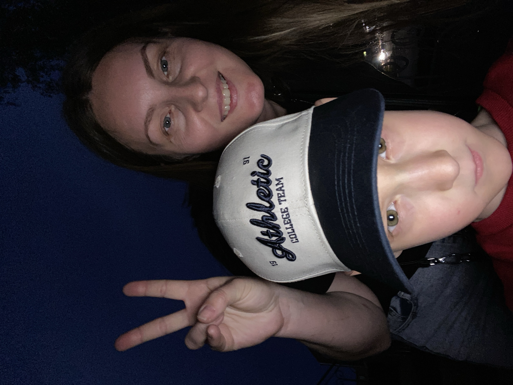 A selfie of a woman and a child, likely taken at dusk or night. The woman is smiling, and the child, wearing a baseball cap, is making a peace sign with their fingers. The cap has the words "Athletic College Team" on it, along with the number "91". The background is dark blue, possibly the night sky. The location within the city of Noviy, Russia cannot be determined from this image.