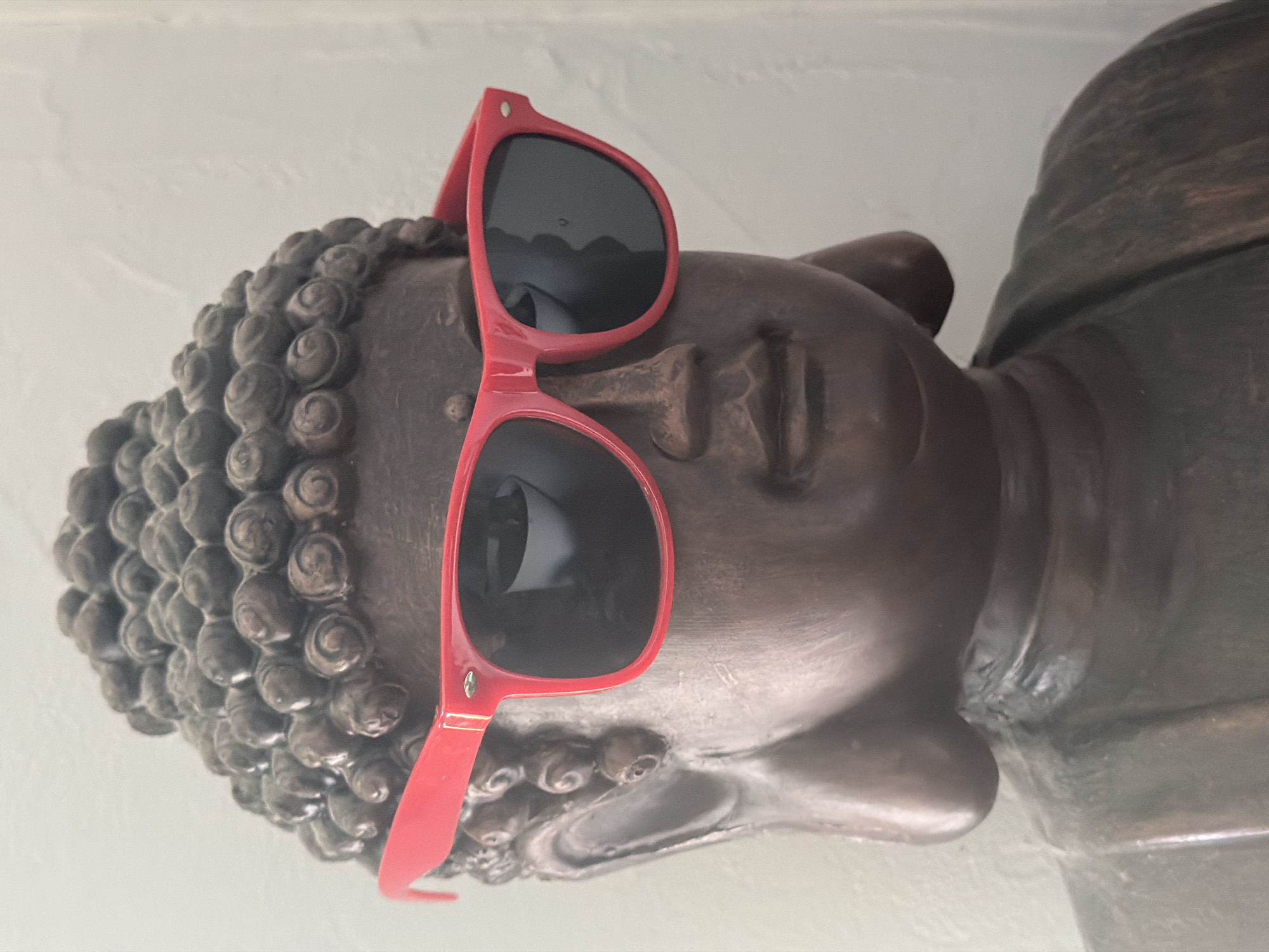 A close-up image depicts a bronze-colored Buddha head wearing red sunglasses. The head features the characteristic tightly curled hair. The sunglasses are positioned on the Buddha's face, obscuring the eyes. The statue is likely indoors. It is unknown if this is located in Esch-sur-Alzette, Luxembourg.