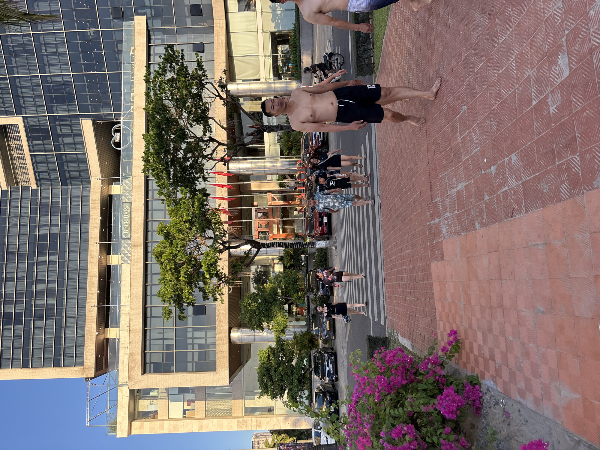 The image shows a view of a hotel in the city of Da Nang, Vietnam, with people walking on a pedestrian crossing and sidewalk in front of it. The hotel is a large, modern building with a facade of glass and beige stone. It has multiple stories, and the windows are covered with blinds. Trees and flowers are visible in front of the hotel.
Several people are crossing the street at a zebra crossing. On the right side of the frame, a bare-chested man in black swim shorts is walking towards the camera. Another bare-chested man in patterned swim shorts is visible in the background to the right. A group of people, including women in dresses and a child, are crossing the street in the middle ground. Further to the left, two women are walking across the pedestrian crossing.
Cars are parked on the street behind the pedestrian crossing. The sidewalk in the foreground is paved with reddish-brown tiles. A bush with bright pink flowers is located on the left side of the sidewalk. The sky is clear and blue, indicating it is daytime. The scene appears to be a public area, possibly near a beach or promenade, given the attire of the people and the presence of palm trees.