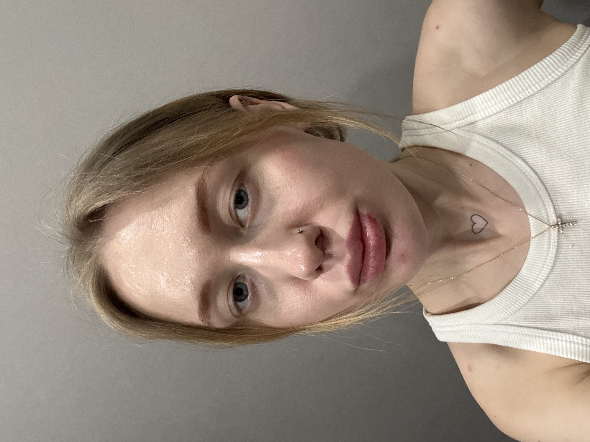 The image presents a close-up portrait of a female individual, situated indoors against a plain, uniform light gray background. The specified location is Rostov-on-Don, Russia. The individual has blonde hair and blue eyes, which are directed forward towards the viewer. A small, metallic stud piercing is observed on the right nostril. Her lips are full, exhibiting a slight puckered expression, with the lower lip displaying a reddish discoloration, particularly towards the right corner. A small reddish blemish is visible inferior to the center of the lower lip. The skin appears fair, with minor blemishes noted on the forehead and chin areas. The individual is wearing a white, ribbed tank top. A thin, gold-colored chain necklace with a small, cross-shaped pendant, adorned with clear stones, is visible around her neck. A distinct black outline tattoo of a heart is located on the right side of her neck. A small, circular red blemish is prominent on her right shoulder, and a fainter blemish is present on her left shoulder.