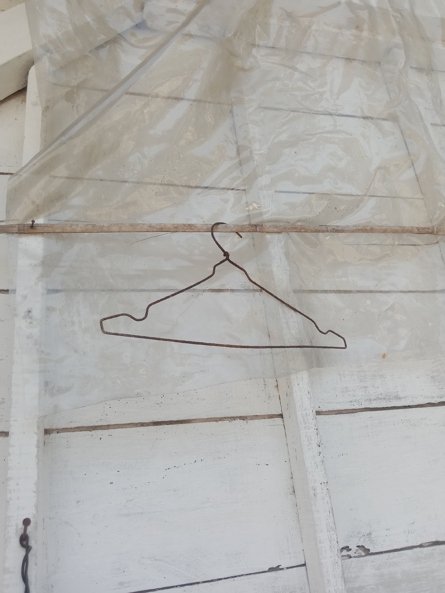 A rusty wire clothes hanger is suspended on a thin wooden rod, which is itself attached to a white wooden wall covered by a translucent plastic sheet. The plastic sheet is wrinkled and translucent, obscuring the details of the wall behind it. The wooden wall is composed of horizontal planks, painted white, with some signs of wear and tear. The scene appears to be outdoors, possibly in a sheltered area or an unfinished construction site in Stiaji, Indonesia. There are no people present in the image.
