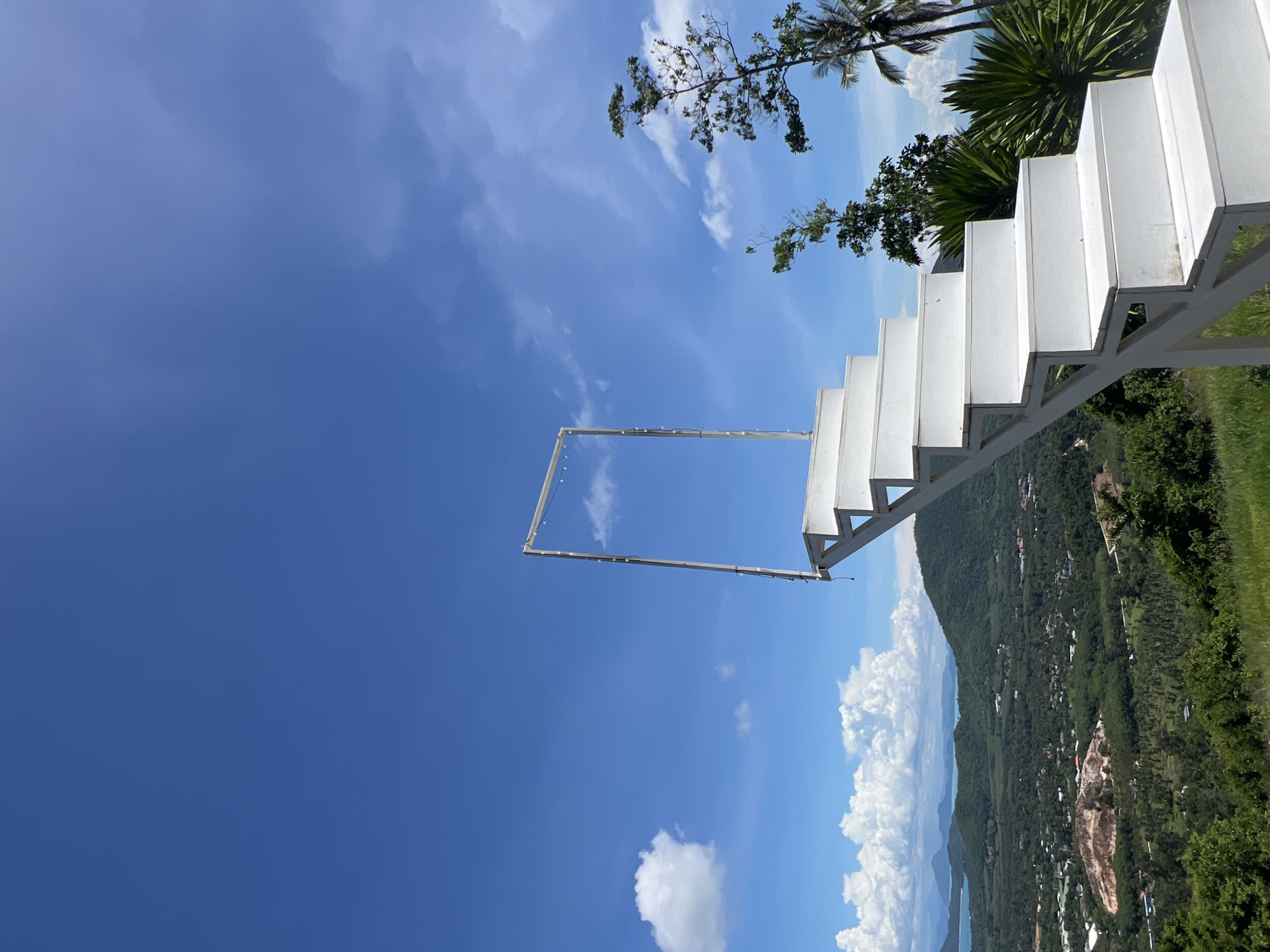 A white staircase leads to an empty frame against a bright blue sky with scattered white clouds. The staircase is positioned on a grassy hill, overlooking a lush green valley with scattered buildings and a body of water in the distance. The scene appears to be in Baan Thurian, Thailand, given the tropical vegetation, including palm trees, visible in the background. The overall atmosphere is serene and picturesque.