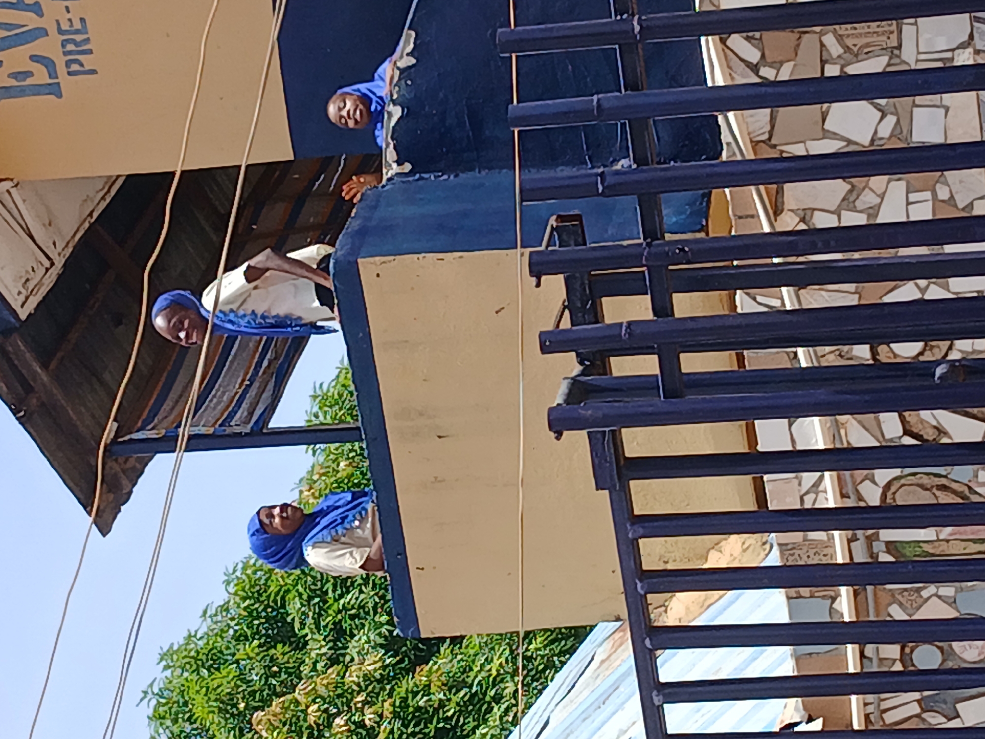 The image, oriented vertically and rotated 90 degrees counter-clockwise, captures an outdoor scene in bright daylight, likely in Badiko, Nigeria. Three individuals, appearing to be women or girls, are visible on a building's upper floor or balcony. All three wear blue hijabs and light-colored tops. Two of them are leaning over a dark blue and yellow/tan painted ledge, looking downwards with smiles, while the third is partially visible further back, looking towards the upper-left of the frame. One individual's top features a blue patterned trim.
A prominent dark metal railing, possibly part of a staircase or fence, occupies the right side of the image. Through its horizontal bars, a wall adorned with a mosaic of irregularly shaped tiles in various shades of white, beige, and brown is visible. Below the railing, a corrugated metal roof can be seen.
In the upper-left portion of the frame, a light-colored sign or banner is partially visible, displaying the vertical text "LIVE" and "PRE-". Above this, a striped metallic canopy or roof is present. The background reveals a clear blue sky and lush green trees in the lower-left corner. Several thin wires or cables hang vertically across the scene. The bright sunlight suggests it is daytime, and the expressions of the individuals indicate a calm and pleasant atmosphere.
