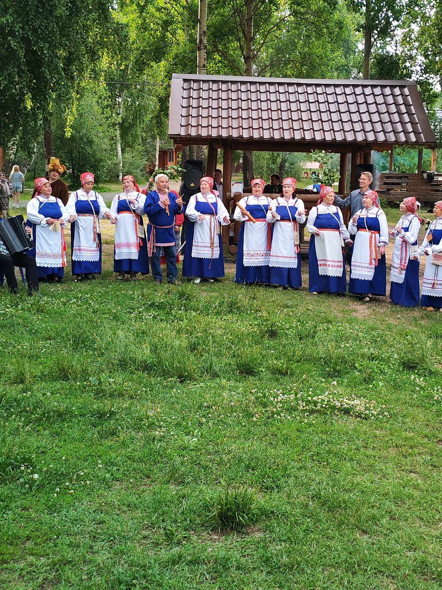 A group of about ten people are gathered outdoors, dressed in traditional ethnic costumes. The group appears to be performing or posing for a photograph in front of a wooden gazebo with a brown tiled roof. The people are standing in a semi-circle on a grassy area, with trees and foliage visible in the background. The costumes consist of long blue dresses with white overskirts and red head coverings for the women, and blue tunics with red trim and pants for the men. One man in the center is holding a microphone, suggesting a performance. An accordion is visible on the left side of the frame, and a speaker is placed behind the gazebo. The overall setting suggests a cultural event or celebration in a park or natural area. This image was captured in the city of Chaikovsky, Russia, likely during a summer day given the lush green foliage and bright lighting.