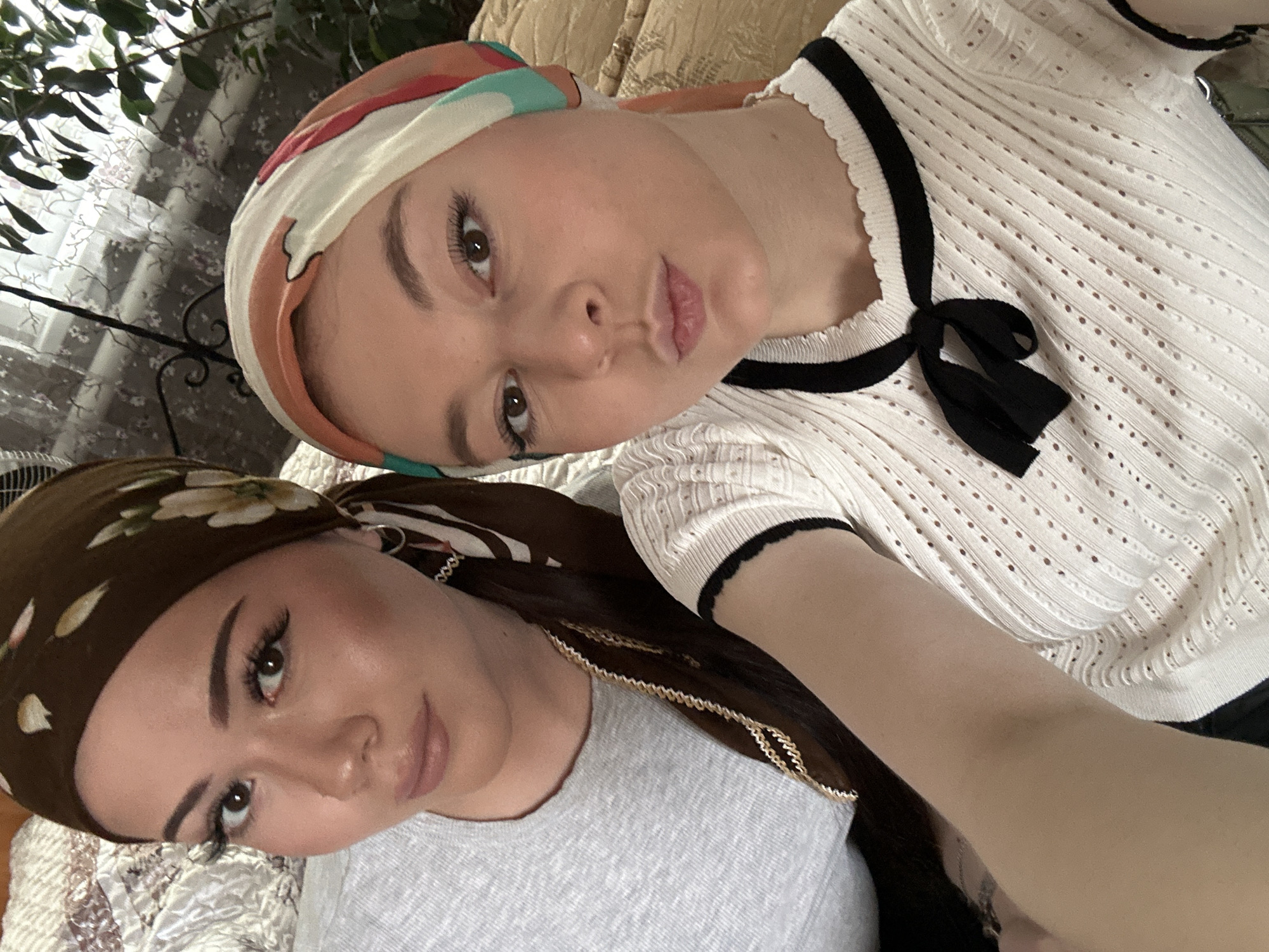 Two women pose closely for a selfie indoors. The woman at the top of the frame wears a white, patterned headscarf and a white, short-sleeved top with black trim and a bow at the neckline. The woman at the bottom of the frame wears a brown headscarf with a floral pattern, a grey t-shirt, and a gold chain. The exact location of the image within Коноваловка, Russia cannot be determined from the image. The environment includes foliage in the background.