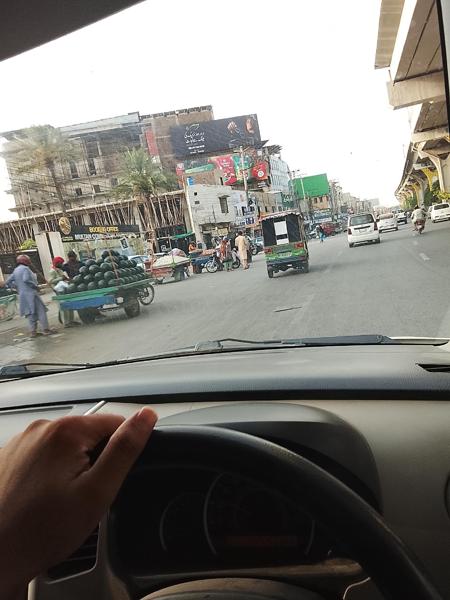 The image shows a view from inside a car driving on a street in Gulgasht Colony, Pakistan. The driver's hand is visible on the steering wheel. The street has several vehicles, including a green auto rickshaw, white vans, and motorcycles. There are people on the sidewalks and crossing the street. A vendor is selling watermelons from a cart on the side of the road. Buildings are visible in the background, some with advertisements and billboards. The sky appears overcast. There are elevated train tracks to the right.