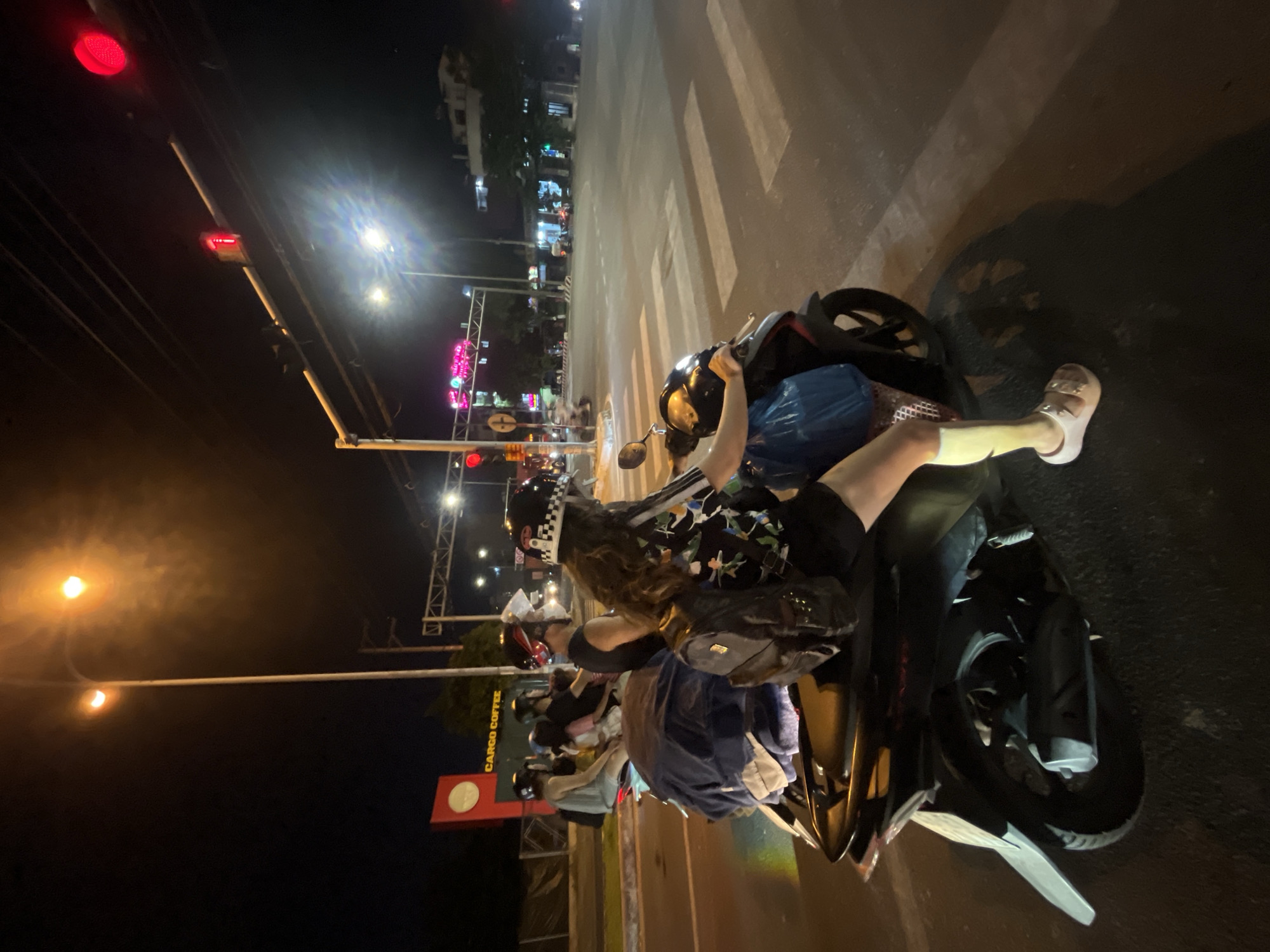 The image depicts a nighttime street scene in Đà Nẵng, Vietnam, with a focus on several people on motorbikes at a street intersection. In the foreground, a person wearing a black shirt with a colorful floral pattern, a black backpack, and white sandals is seated on a black motorbike, facing away from the camera. Their right leg is extended, resting on the motorbike's body. The motorbike is loaded with bundles of clothing and a blue plastic bag. Behind this individual, another motorbike carries a passenger wearing a black helmet with a checkered pattern.
Further back, other individuals are visible on motorbikes. One person in a gray tank top is on a motorbike in the background. A red sign with a white circle and the text "CARGO COFFEE" is visible on the left side of the image, suggesting the presence of a coffee shop or establishment. Traffic lights are illuminated, with a red light visible in the upper right portion of the image, indicating the intersection is active. The road surface includes a pedestrian crossing. The overall environment is urban, with streetlights and building lights illuminating the night. The scene captures a moment of transit or waiting at a traffic signal.
