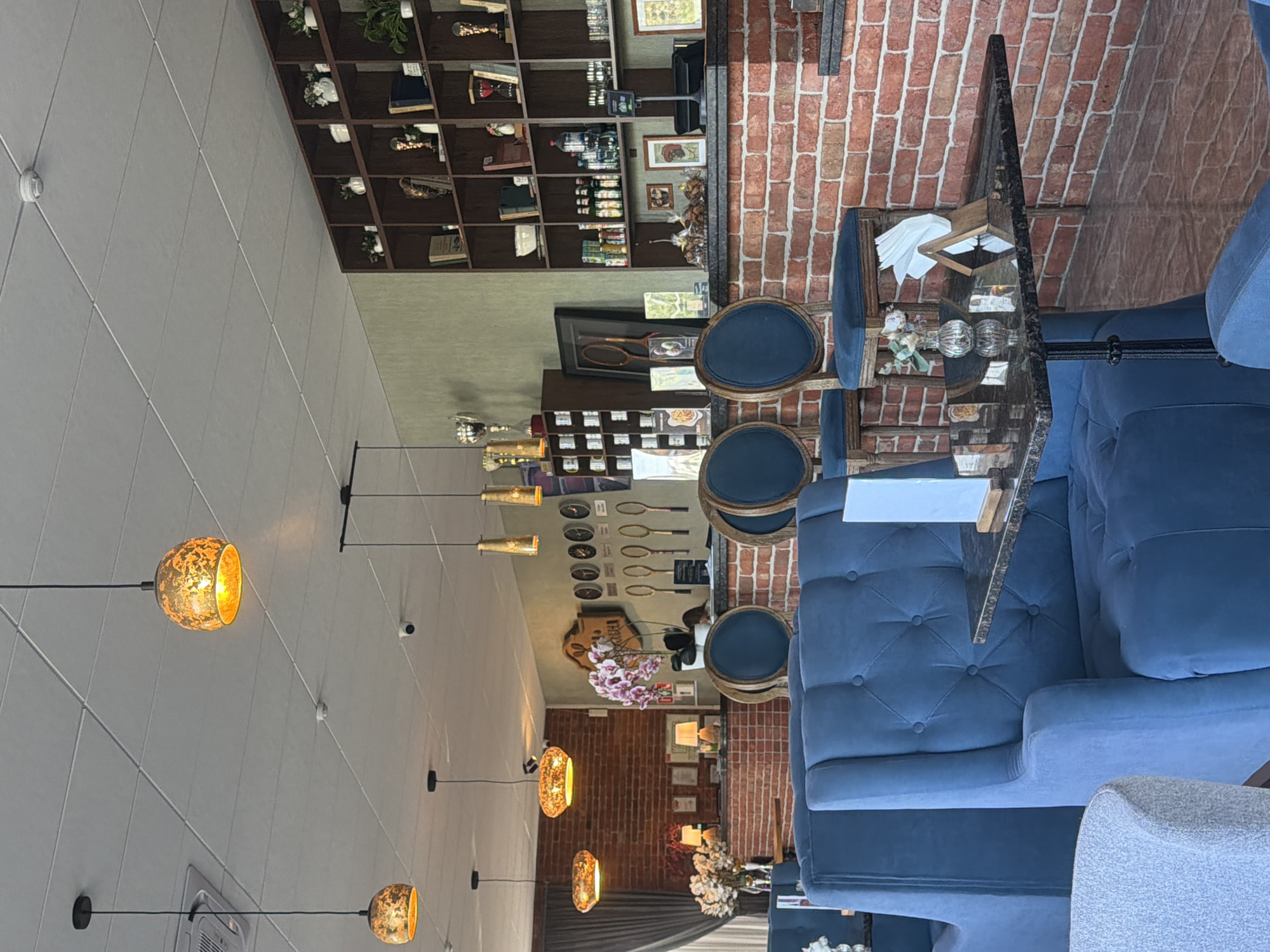The image depicts the interior of a restaurant or cafe. The space features a combination of design elements, including a wall of brickwork and shelving units displaying decorative items. Tables are set with blue seating and glass tops. Multiple pendant lights with a golden hue are suspended from the ceiling, providing illumination. There is no evidence to indicate that this is in the city of Ovoshchnoy, Russia.
