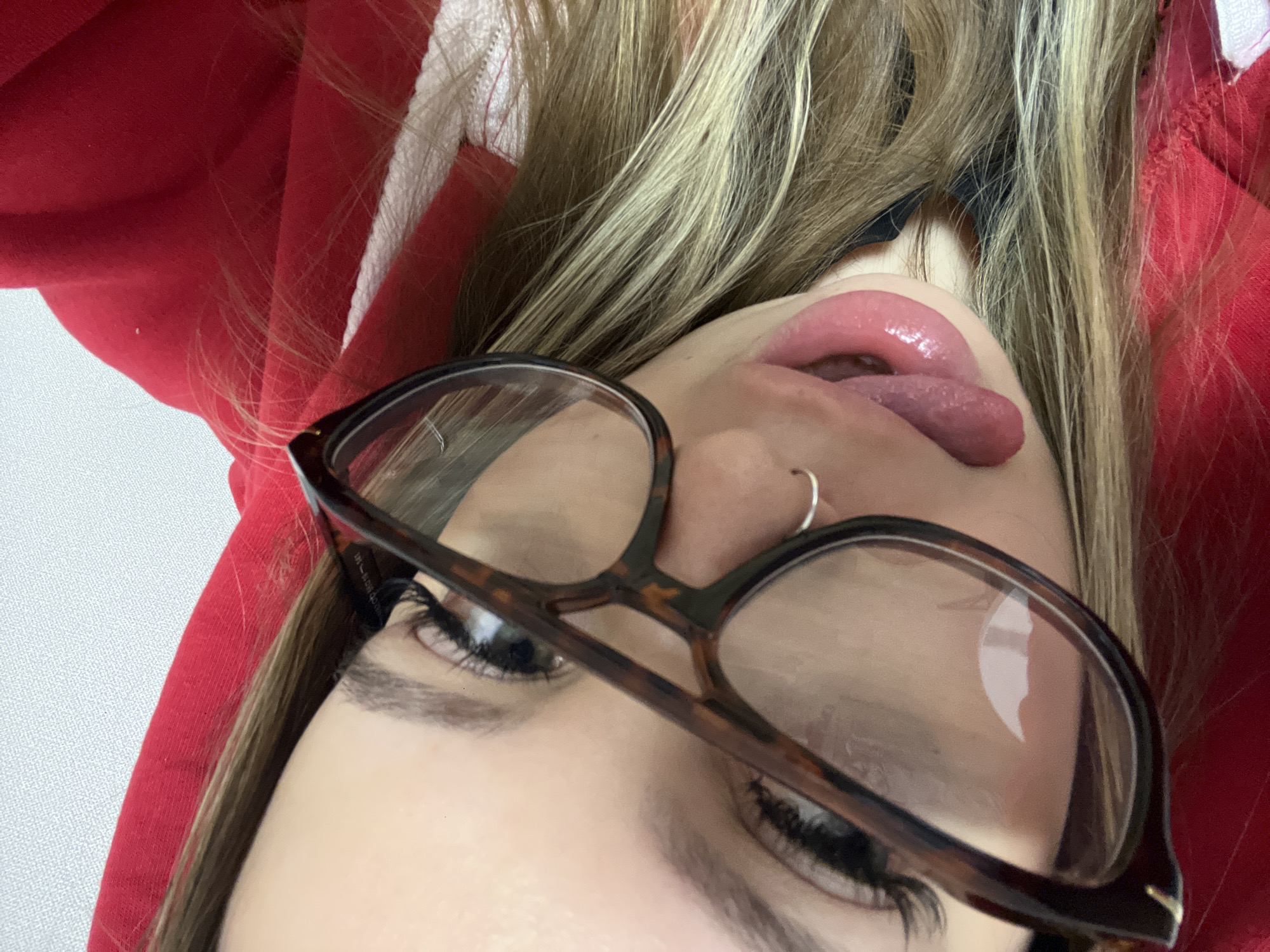 A close-up, tilted selfie depicts a woman with blonde hair and glasses sticking out her tongue. She wears a red hooded sweatshirt. A silver nose ring is visible. The background is indistinct and does not provide enough information to determine if it is in the city of Krasnodar, Russia.