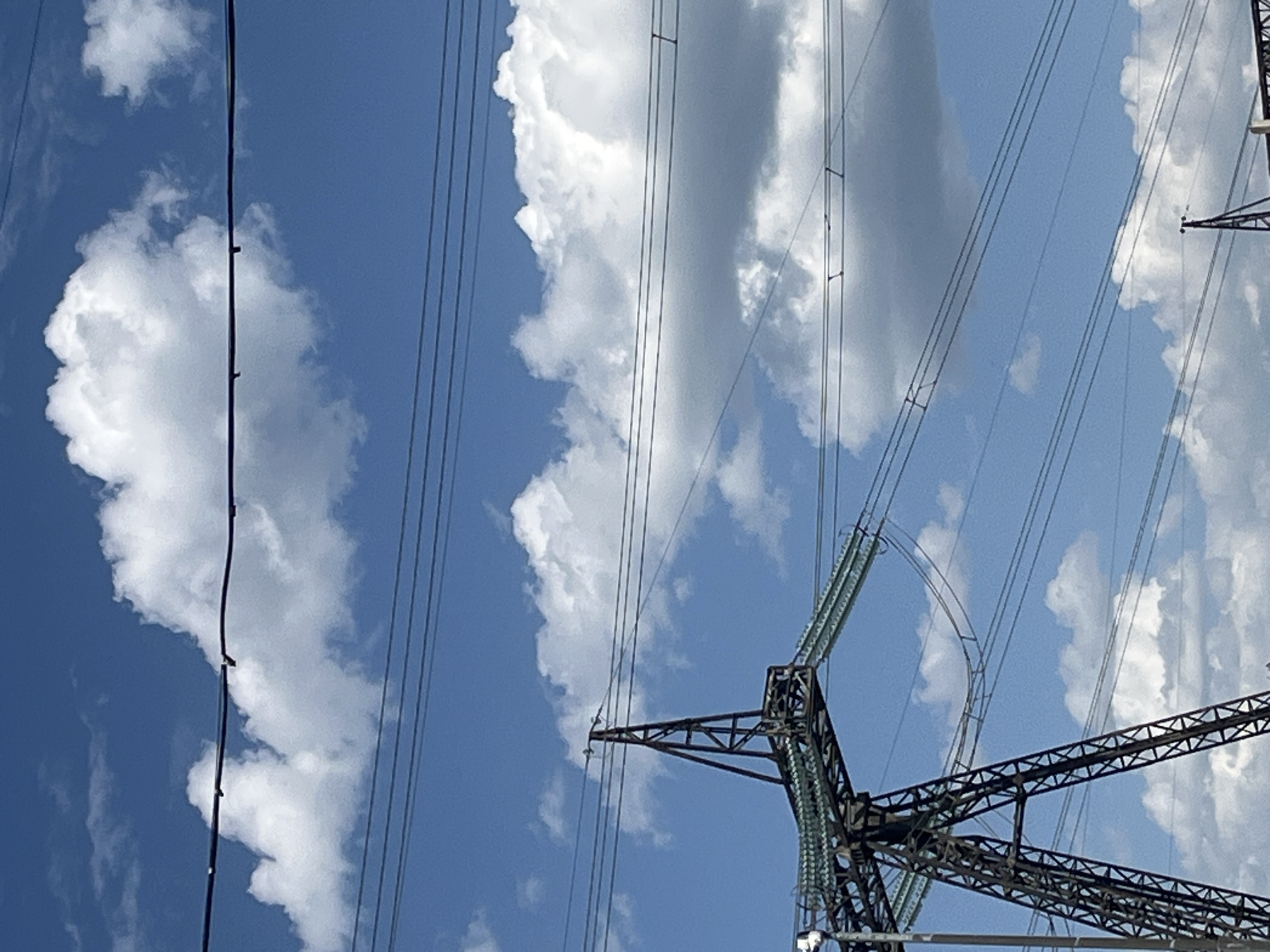 The image depicts a view looking up at power lines and a metal power transmission tower against a partly cloudy blue sky. The lines are suspended from the tower and stretch diagonally across the frame. Cumulus clouds are visible in the background. The location in Chaikovsky, Russia, cannot be verified from the visual content of the image alone.