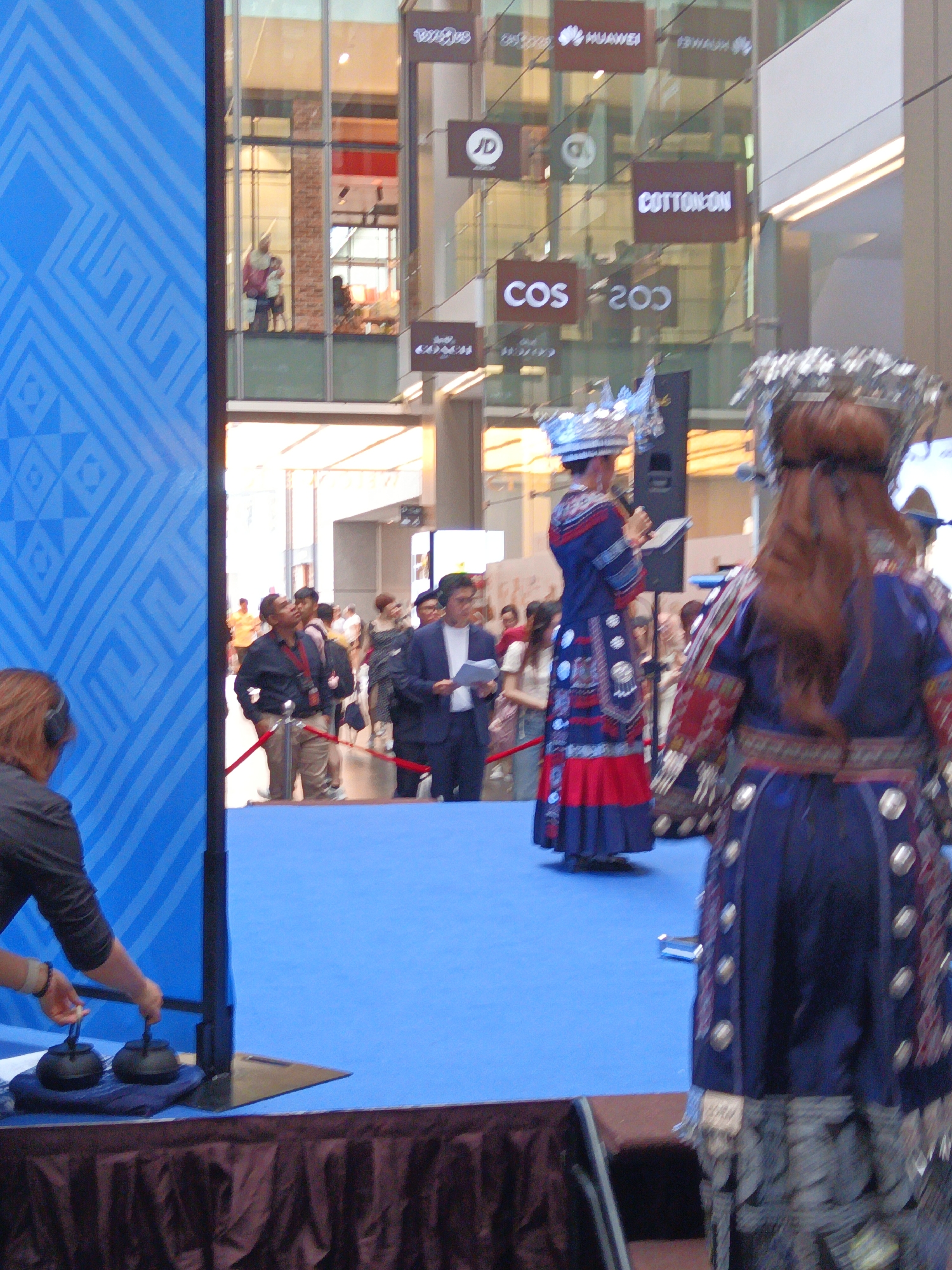 The image depicts an indoor event, possibly a cultural performance or exhibition, taking place at Suria KLCC in Kuala Lumpur, Malaysia. In the foreground, a person wearing headphones and a dark top is partially visible, engaged with two small black teapots placed on a navy blue mat. To the right, two individuals in elaborate traditional blue and red attire, adorned with intricate silver embellishments and headdresses, are the central focus. One is speaking into a microphone, standing on a bright blue carpeted stage. Behind them, a crowd of onlookers and participants are visible, some in formal wear and others in traditional clothing. The background reveals a multi-story glass-fronted building with various retail store signs, including "HUAWEI," "JD," "COTTON ON," and "COS," indicating a shopping mall environment. The lighting suggests an indoor, well-lit space.