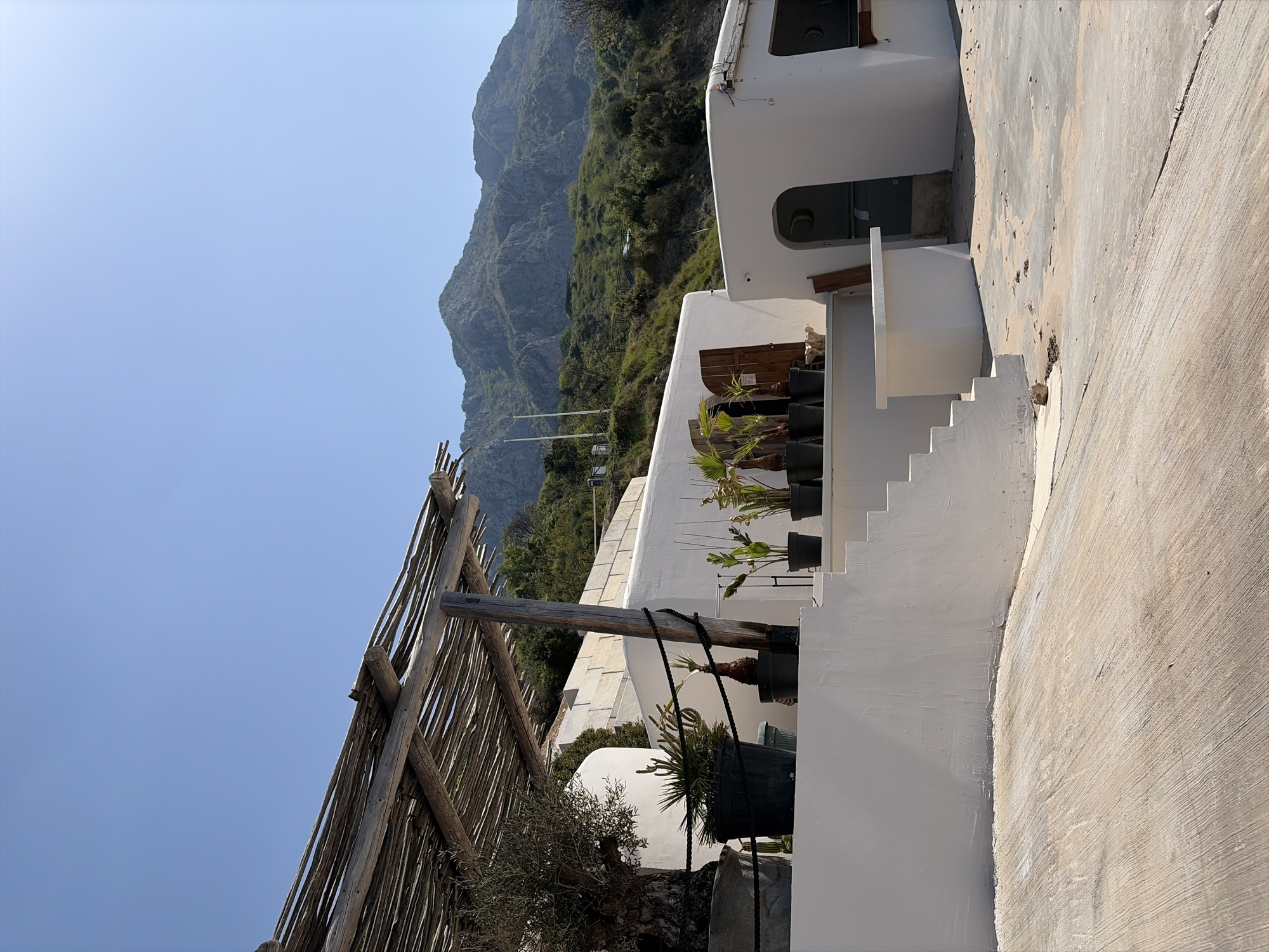 The image displays a brightly lit outdoor scene, originally oriented vertically, showcasing a complex of white buildings nestled against a large, verdant mountain under a clear blue sky.
On the right side of the frame, several levels of white, stucco-style buildings are visible, characterized by minimalist architecture. These structures incorporate multiple outdoor staircases and terraces, some of which are adorned with numerous plants in black pots, including small palm trees and other foliage, providing touches of greenery. A light-colored, possibly concrete, paved pathway or terrace is prominent in the foreground and extends throughout parts of the complex.
Dominating the left side and background is a majestic, rugged mountain covered in lush green vegetation and rocky outcrops. On the far left, a rustic wooden fence or trellis, constructed from natural, rough-hewn branches, partially frames the scene, hinting at an outdoor area.
The scene is bathed in strong, natural sunlight, evident from the distinct shadows cast by the buildings and foliage, and the vibrant, cloudless blue sky, indicating a clear day, likely midday or early afternoon. The architectural style and mountainous landscape evoke a serene Mediterranean or island setting. No human activity or visible text is present in the image.