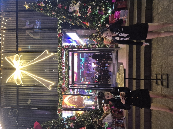 The scene depicts the nighttime exterior of a commercial building entrance in Thuận Hóa Ward, Vietnam, extensively decorated for the Christmas season. A large, illuminated bow-shaped light fixture is prominently centered above the glass-paneled facade. Green garlands adorned with lights and "MERRY CHRISTMAS" text frame the open doorway. Two decorated Christmas trees flank the entrance, each embellished with lights, various ornaments, and wrapped gift boxes at their bases; a reindeer figurine is visible near the left tree. The open glass doors reveal a brightly lit, colorful interior, resembling a bar or club, with multiple patrons seated at tables. A set of shallow steps leads from the paved ground level to the entrance. Two women stand on the paved area outside the steps. The woman on the left, dressed in a black uniform-style blazer with gold buttons, a white patterned tie, and a black skirt, is holding a smartphone with both hands and looking down at its screen. The woman on the right, wearing a black long-sleeved dress and a white sash bearing "J'ADIOR" text, is adjusting her left shoulder or neck area with her left hand while holding a light-colored object in her right hand. A black metal stand displaying several gold-colored plaques is positioned on the ground between the two women.