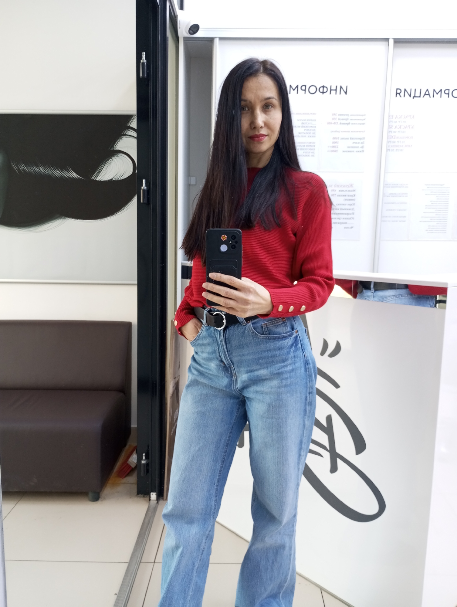 A woman is taking a selfie in what appears to be a commercial space, possibly a store, with the city of Izhevsk, Russia identified as the location. The woman is wearing a red long-sleeved sweater, blue jeans with a black belt, and her dark hair is long and straight. She is holding a black smartphone in her right hand to take the picture. She has one hand in her jeans pocket.
Behind her is a reception area with text written in Cyrillic. A brown sofa is visible to her left. A security camera is mounted on the upper wall above her. The setting looks like a modern, well-lit interior with a tiled floor.