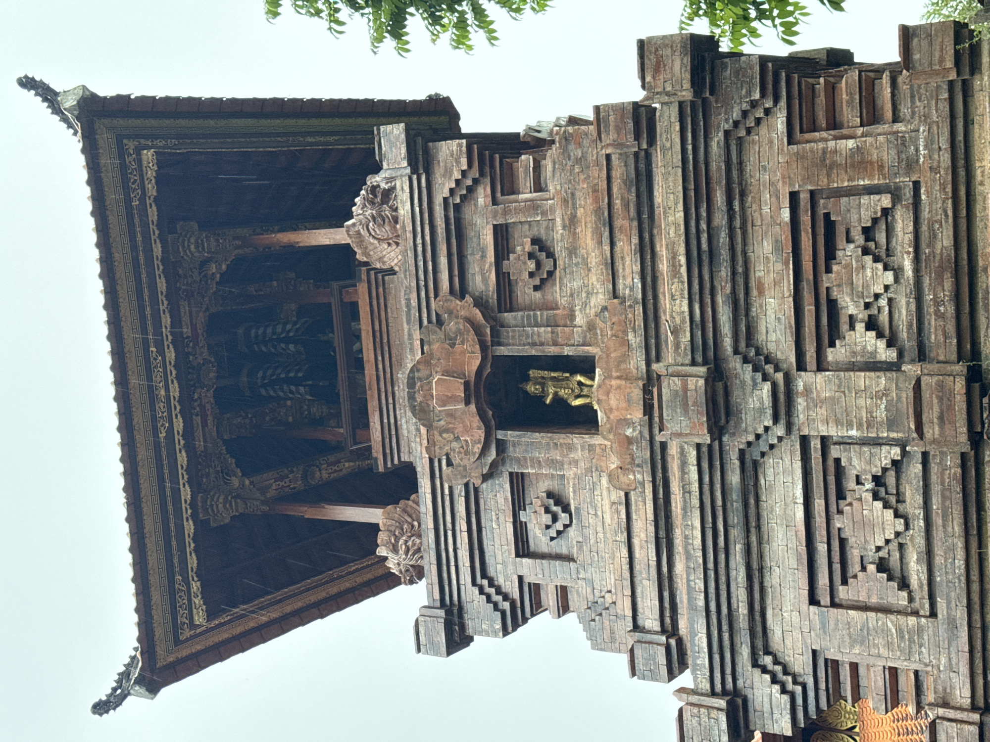 An elaborate, multi-tiered architectural structure, likely a traditional Balinese temple gateway or building, is depicted from a low-angle perspective against a bright, overcast sky. The structure features a facade constructed from reddish-brown brick or carved stone, characterized by intricate stepped patterns and geometric motifs. Within a central niche on the facade, a small, golden statue of a deity or religious figure is prominently placed.
The upper portion of the building showcases a dark wooden roof with richly decorated eaves. These eaves are adorned with gilded or golden-colored patterns and elaborate wood carvings, some of which appear to be stylized mythological figures or floral designs. Supporting beams with detailed carvings are also visible beneath the roof. Green foliage from a tree is visible at the very top edge of the frame, providing a natural accent.
No human figures or explicit activities are taking place; the image is a static portrayal of the architecture. The bright, uniform sky suggests a daytime setting, possibly under overcast conditions. The architectural style, with its ornate carvings, tiered design, and material usage, strongly indicates a location in Bali, Indonesia, consistent with Kuta Selatan. There is no visible text in the image.