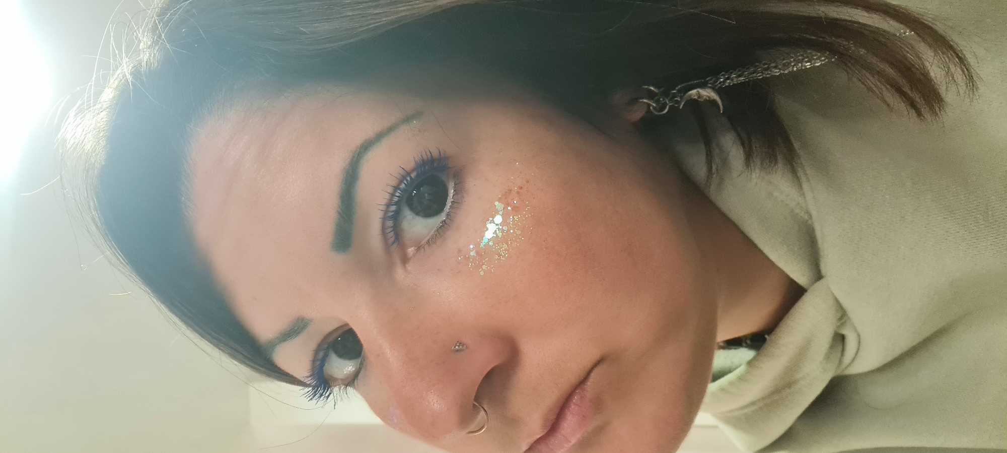 A close-up, slightly angled shot of a person's face. They have dark hair, blue mascara, and glitter near their eye. There is a nose piercing, a ring on their nostril, and what appears to be a silver crescent moon earring. The person wears a light-colored hooded sweatshirt. The location and action cannot be determined from the image.