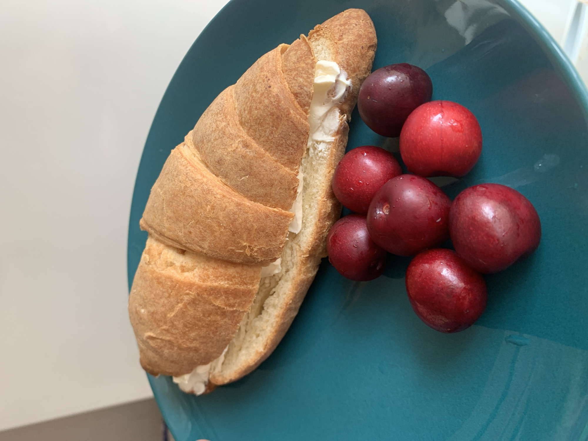 A single croissant filled with white cream cheese is presented on a teal plate. Beside the croissant, a cluster of seven red cherries is arranged. The scene is captured from a top-down perspective with no people visible. The background is a plain white wall. The location is Kapyĺ, Belarus.