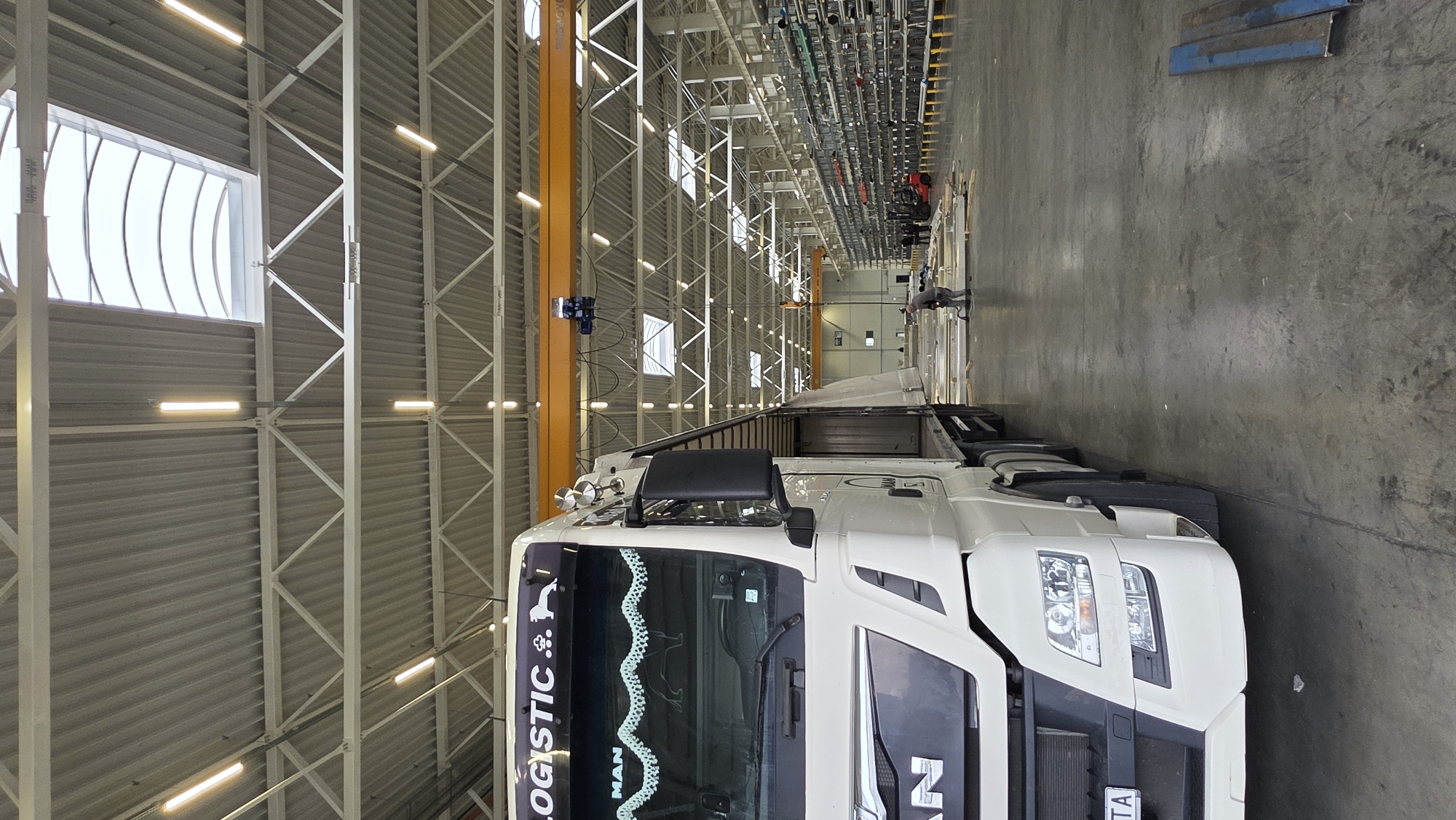 A white MAN truck is parked inside a large industrial building in Gara Elin Pelin, Bulgaria. The building features a high ceiling with exposed steel beams and artificial lighting. An overhead crane runs along the length of the building. A person is standing near the back of the truck, possibly overseeing the loading or unloading process. The concrete floor is a uniform gray. Some metal components or materials are visible in the background.