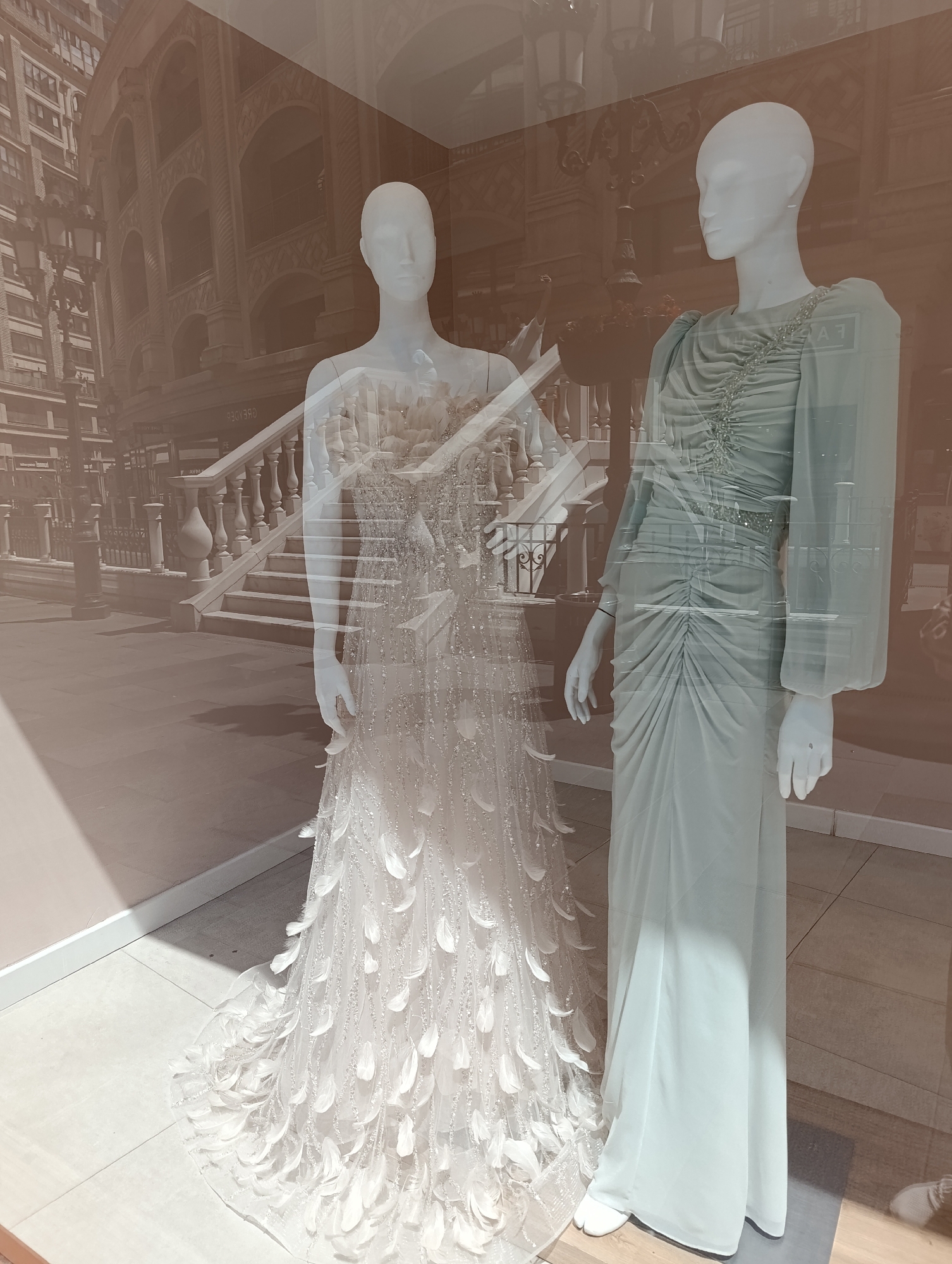 The image shows two mannequins in a shop window. The mannequin on the left is wearing a white, floor-length gown adorned with feathers and sequins. The mannequin on the right is wearing a pale blue, floor-length gown with ruching down the front and long, flowing sleeves. Both mannequins are white and feature smooth, featureless heads. Behind the mannequins, a large photograph or mural is visible, depicting a grand staircase and arched architecture, suggesting an outdoor or historical setting. The reflection of the surrounding environment, including buildings and streetlights, is visible on the shop window. The floor of the shop appears to be tiled. The overall impression is that of a boutique selling formal attire, potentially for special occasions.
The image does not provide any specific information or visual cues that would allow for the identification of its location within the city of Gaziosmanpaşa, Turkey.
