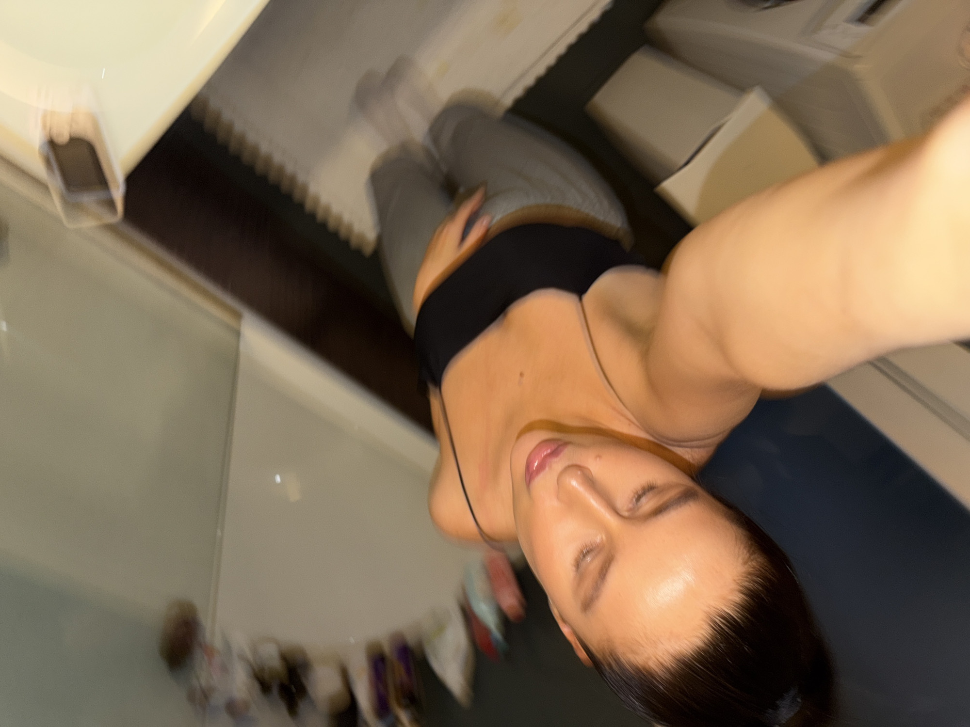 This blurry, angled selfie captures a young woman indoors, likely in a bathroom reflected in a mirror. The primary subject is a woman with dark hair pulled back, wearing a black spaghetti strap top and light grey sweatpants. She looks downwards with her eyes slightly closed, conveying a relaxed and serene expression. Her left hand rests gently on her lower abdomen.
The background elements are largely blurred due to motion or low light. In the upper left, a white surface features a soft, rectangular object that could be a phone or smart watch. Further back, a dark blue or grey wall or floor contrasts with white fixtures and reflective surfaces. Various indistinct objects, possibly bottles or toiletries, are visible on shelves or counters, reflected in the background. On the right side, a portion of a white appliance, resembling a washing machine, can be seen. A distinctive white, serrated edge runs horizontally across the upper part of the background, possibly belonging to a bathtub or shower curtain track. The lighting appears artificial and somewhat dim, highlighting the reflective surfaces and her skin. The image is significantly blurred, suggesting movement during the capture. There is no visible text. The location context provided is Khimki, Russia.
