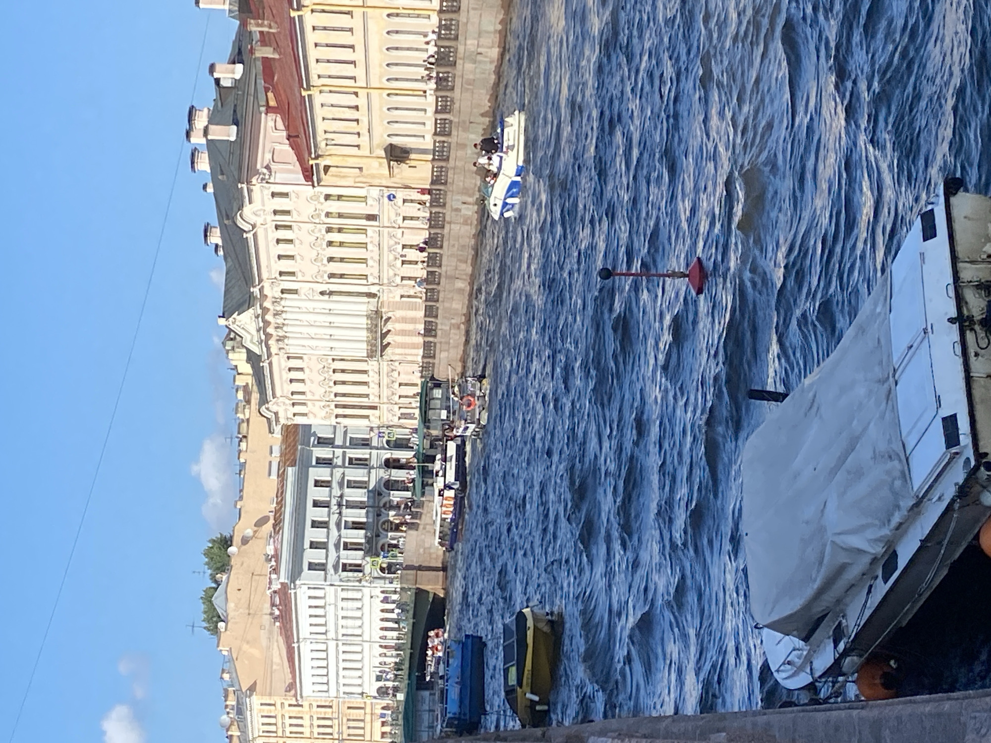 The image captures a sunny day with a bright blue sky and scattered white clouds over the city of Saint Petersburg, Russia. The foreground is dominated by a wide canal, its surface rippled by a strong current. Several boats are navigating the canal.
In the lower left corner, a white boat is docked, partially covered by a light-colored tarp. Two bright orange fenders are attached to its side.
Further into the canal, a yellow boat with a dark roof is moving away from the viewer. To its left, a dark blue barge is also visible.
In the mid-ground, two passenger tour boats are traveling side-by-side. One is white with a dark roof, and the other is white with a lighter roof. Another smaller boat with a dark roof and a light-colored upper section is also present.
In the right mid-ground, a small white and blue speed boat with four occupants, three visible, is making its way down the canal. A red buoy with a red marker on top is situated in the water between the yellow boat and the speed boat.
The background features a row of ornate, multi-story buildings lining the canal's edge. These buildings exhibit classical architectural styles with arched windows and detailed facades, painted in various shades of cream, yellow, and pale pink. A pedestrian walkway with railings is visible along the canal bank in front of the buildings, with numerous people strolling. A stone bridge arches over the canal further in the distance, with more buildings visible beyond it. The overall scene depicts a vibrant urban waterway on a clear day.