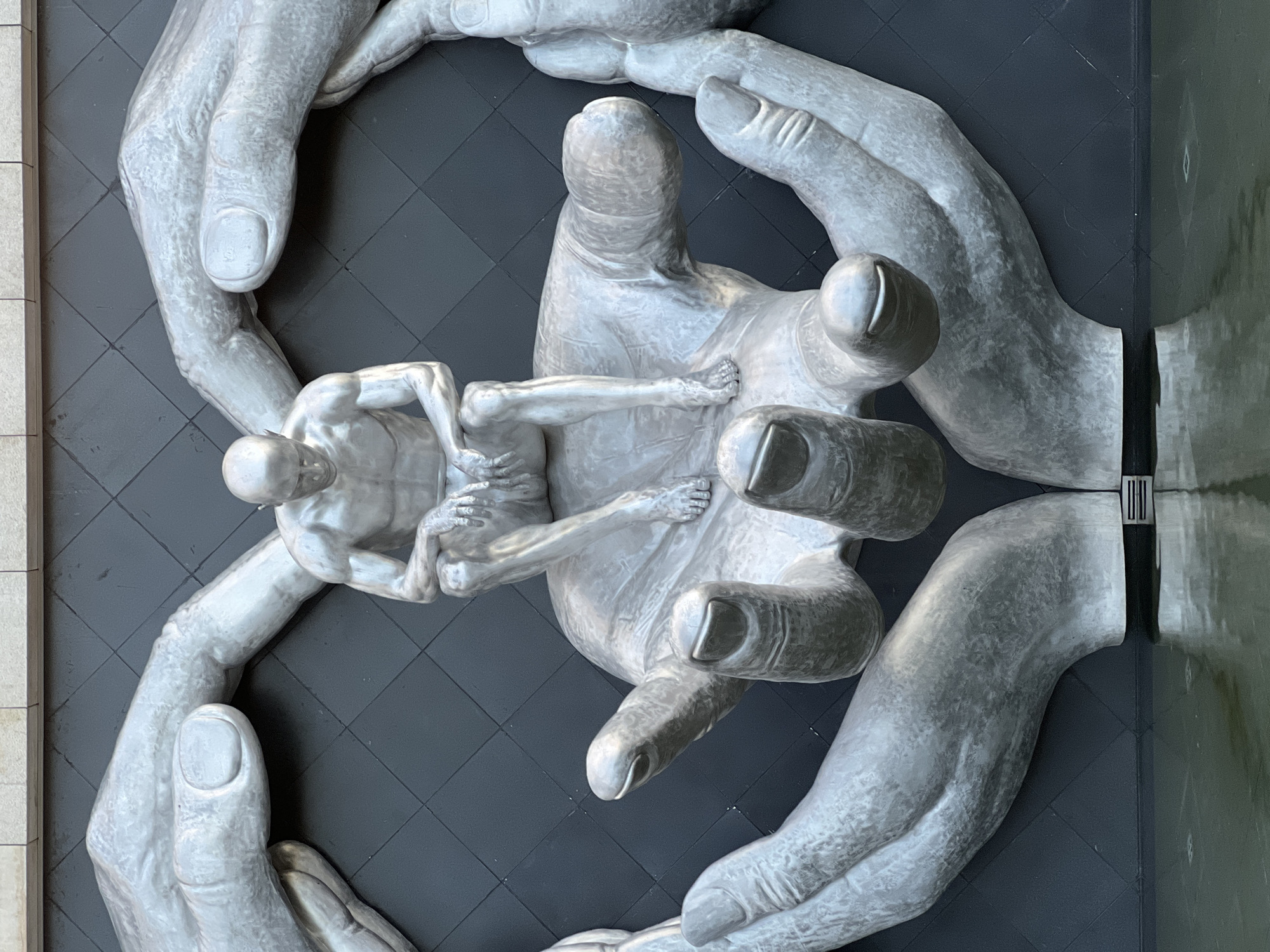A silver statue of a seated man is cradled by multiple oversized silver hands, forming a heart shape. The sculpture is set against a dark, grid-patterned wall, with a pool of water reflecting the artwork in the foreground. The statue depicts a male figure with a bald head, muscular physique, and his arms resting on his knees. The hands are depicted in various positions, some cupping the figure, others forming the outer frame. The setting appears to be an outdoor public space, possibly a plaza or park, given the water feature and the architectural elements visible in the background. The image does not provide information about the specific city or location of Krasnodar, Russia.