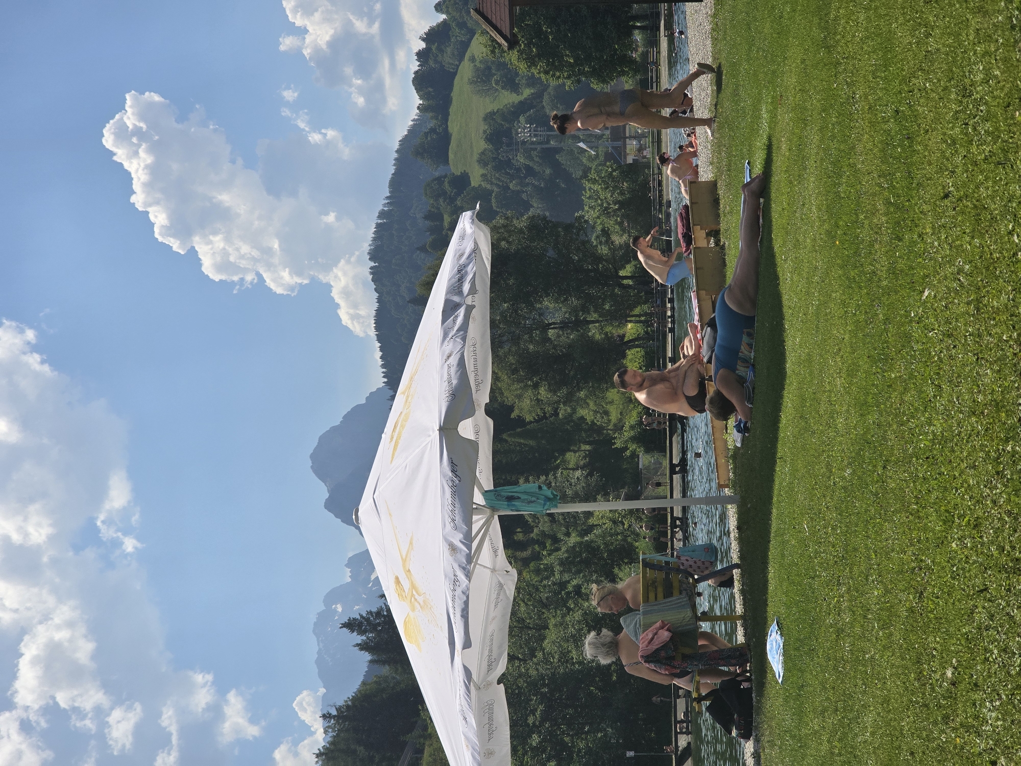 The image depicts a sunny day at an outdoor swimming pool or lido, likely in Garmisch-Partenkirchen, Germany, given the mountainous backdrop. In the foreground, a person lies prone on the grass, sunbathing. To the left, under a large white umbrella adorned with a golden graphic and text, two women sit on a bench. A man is visible to the right of the umbrella, also sunbathing, and further to the right, a woman in a bikini walks away from the camera. The background features a body of water with several people swimming or wading, a wooden deck or pier, lush green trees, and rolling hills leading up to imposing, jagged mountains under a bright blue sky with scattered white clouds.