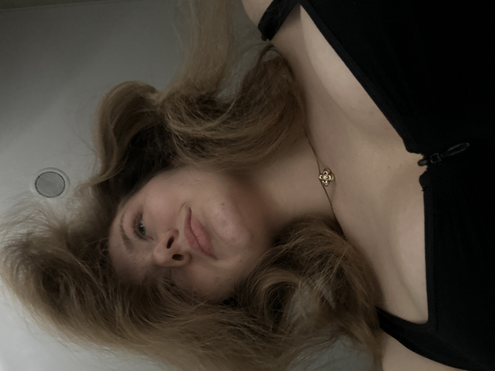 The image shows a woman with blonde, wavy hair and fair skin, lying down and looking at the camera. She is wearing a black top and a gold necklace with a clover-shaped pendant. The background appears to be a white surface, possibly a bathtub or a similar fixture. There is a circular drain visible in the upper left corner of the frame. The location is unknown. There is no information to suggest that the image was taken in the city of Kazan, Russia.