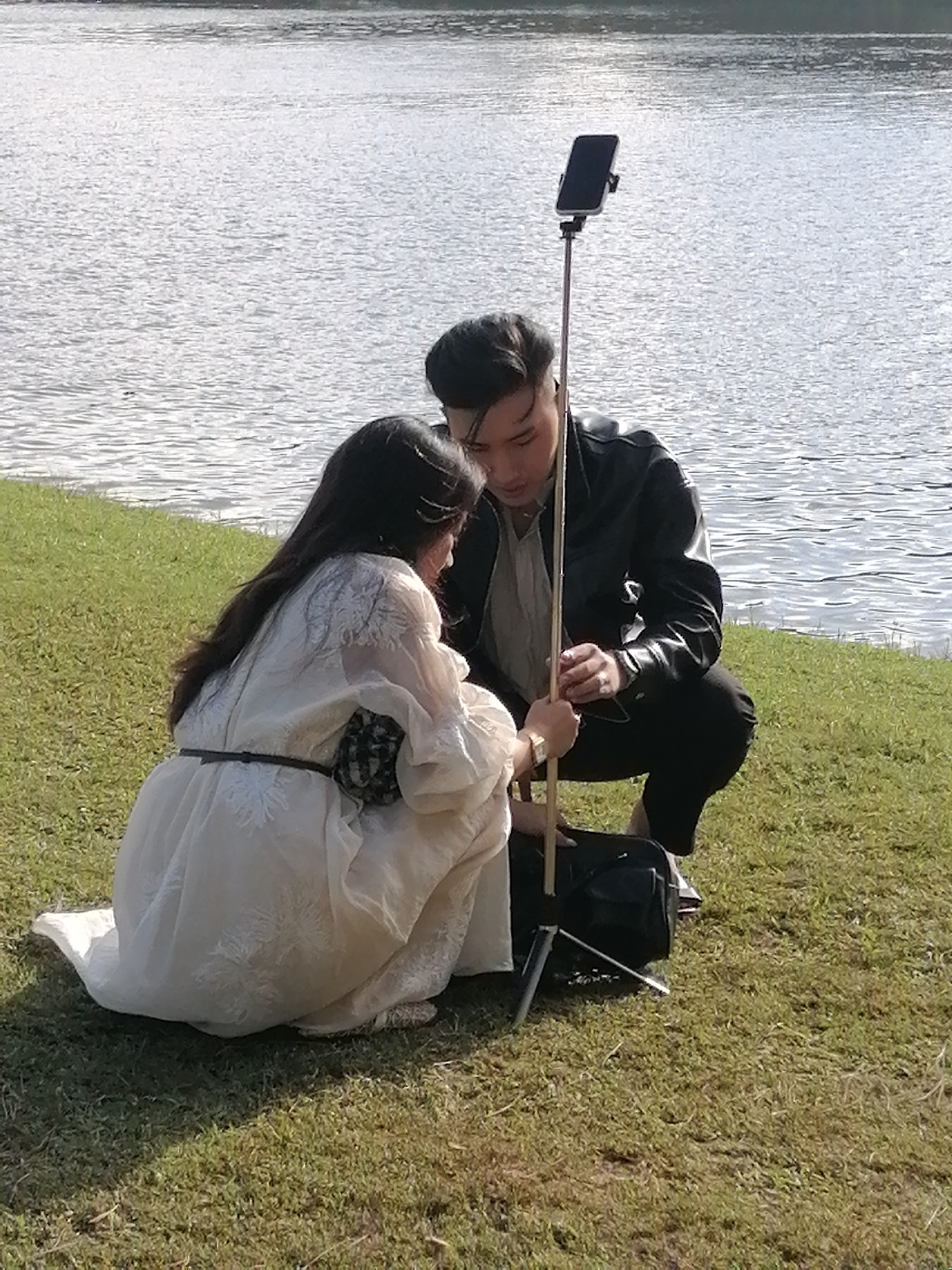 A young Asian couple is posing for a selfie in front of a lake. The woman is kneeling on the grass in a white dress, and the man is crouching beside her, holding a selfie stick with a phone attached. The lake is in the background, and the sun is shining. The scene appears to be in Da Lat, Vietnam, a city known for its natural beauty and lakes.