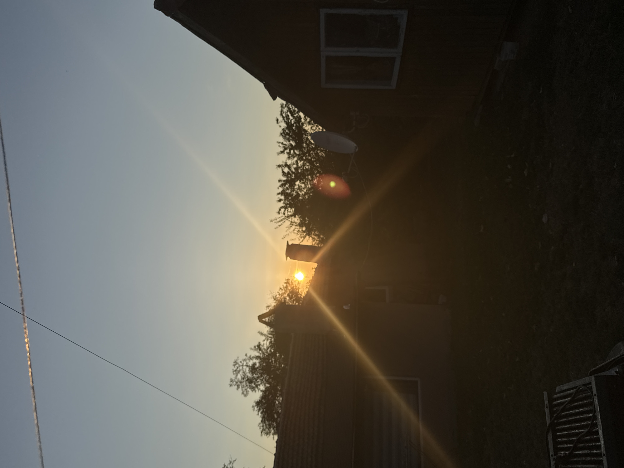 The image depicts a sunset scene with two houses and trees in the foreground, set against a clear sky with a thin contrail from an aircraft. The sun is low on the horizon, casting a warm, golden light that creates lens flares and long shadows. The first house, to the left, has a tiled roof and a chimney. The second house, to the right, is darker and has a pitched roof with a window and two satellite dishes mounted on its side. In the lower left corner of the frame, a grill grate is visible. The ground appears to be covered in grass and fallen leaves. The overall atmosphere is peaceful and serene. The image was taken in the city of Sredets, Bulgaria.