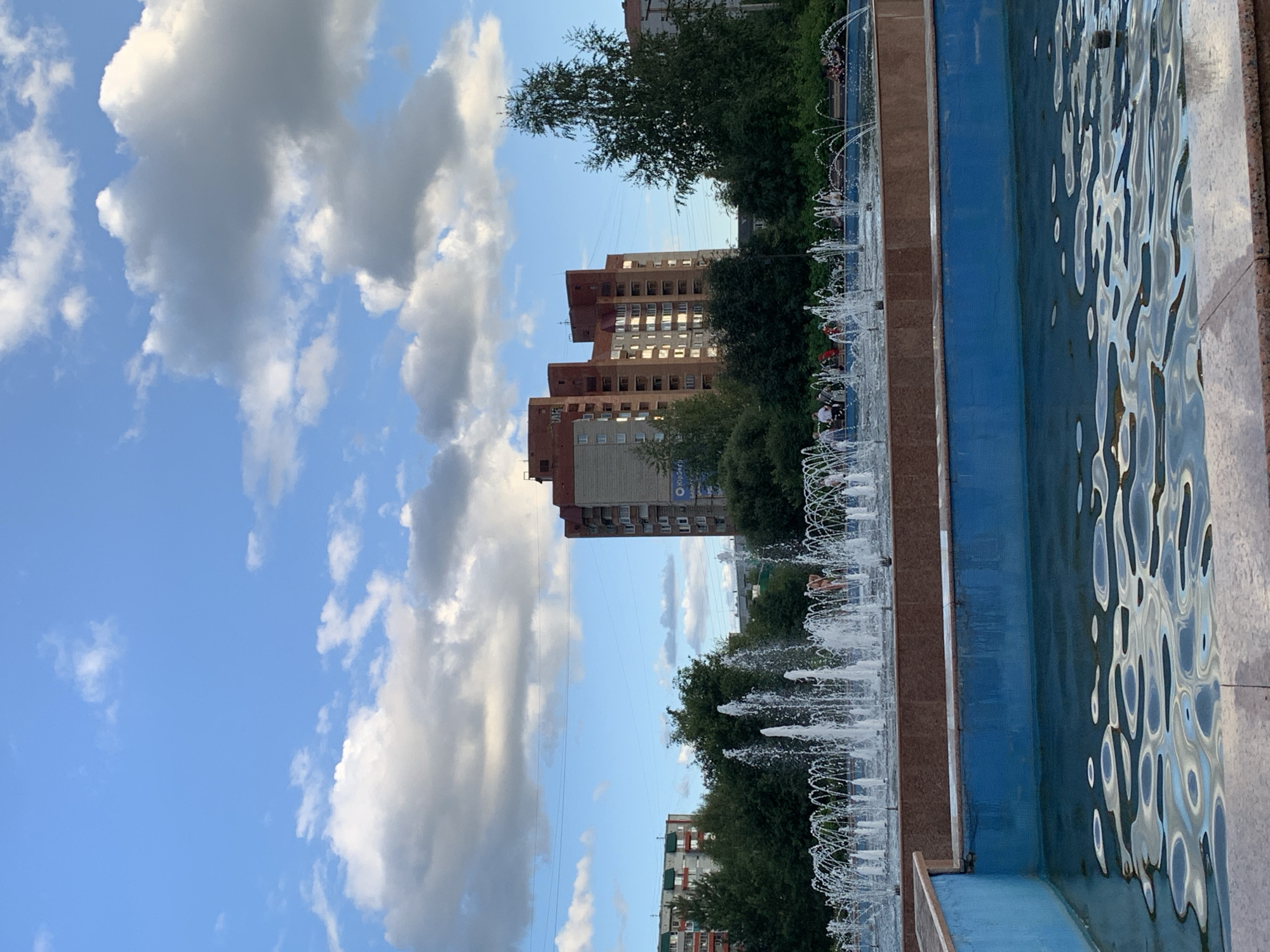 The image shows a city park in Prokopyevsk, Russia, featuring a large fountain with numerous water jets. In the foreground, a blue-tiled pool reflects the sky and surrounding elements. The middle ground shows the active fountain and a raised, reddish-brown tiled area behind it, with people gathered on benches and walking. Beyond the fountain are lush green trees and several multi-story buildings, including a prominent brick-colored apartment building with numerous windows. The sky is a clear blue with scattered white and gray clouds, suggesting it is daytime. Power lines are visible stretching across the sky. The overall scene depicts a public recreational space on a pleasant day.