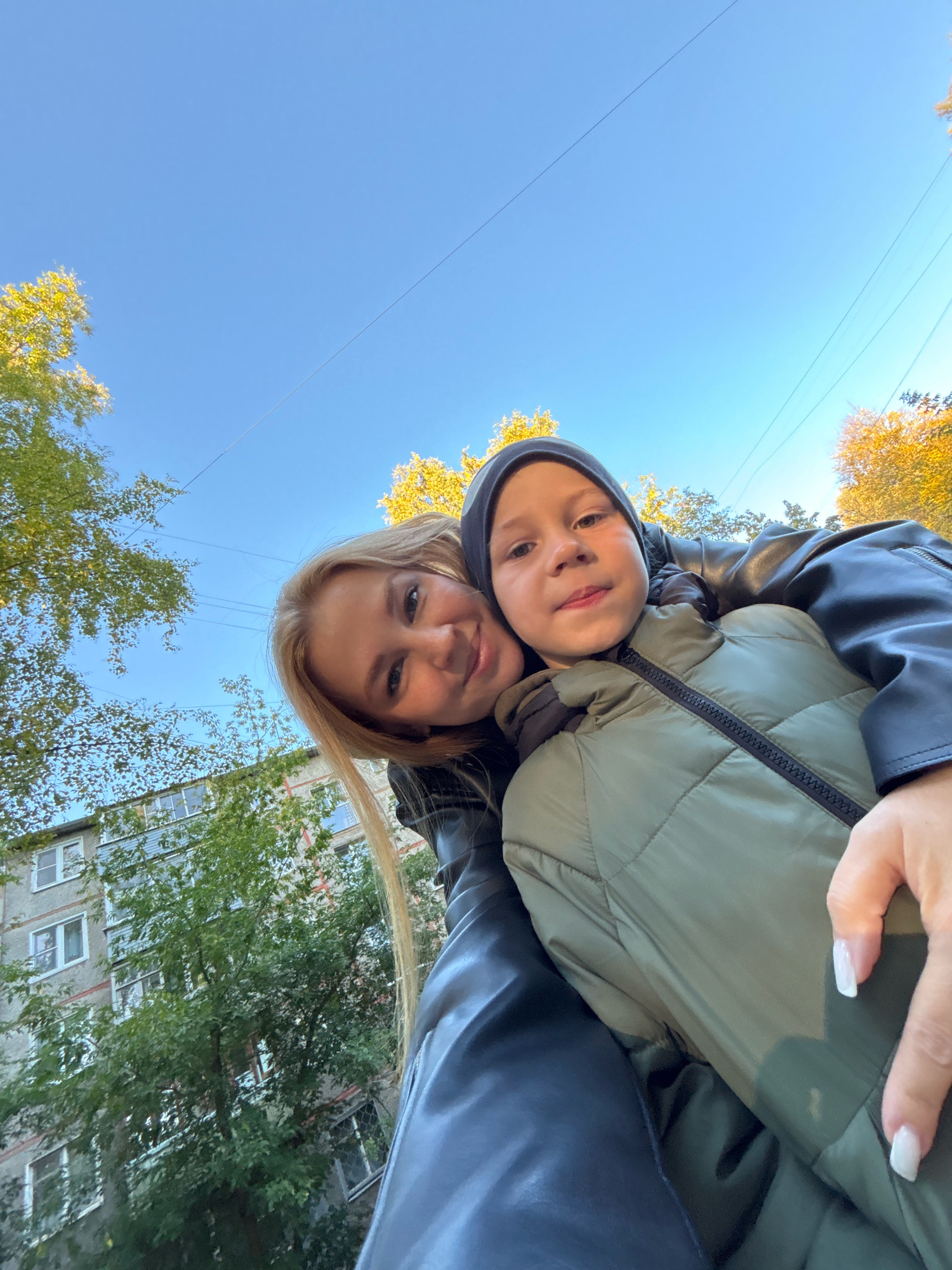 In a low-angle, close-up selfie, a woman with blonde hair smiles widely, her head tilted to her left and her face close to a young boy's. The boy, wearing a dark beanie and a green puffer jacket, also faces the camera, with his tongue slightly out. They appear to be outdoors in Rusanovo, Russia, indicated by a building with visible windows and balconies in the background, surrounded by trees with green and yellow foliage. Power lines are visible against a clear blue sky, suggesting daytime. The woman's hand, with white-polished fingernails, rests on the boy's jacket. Both are wearing outerwear, indicating cooler weather.