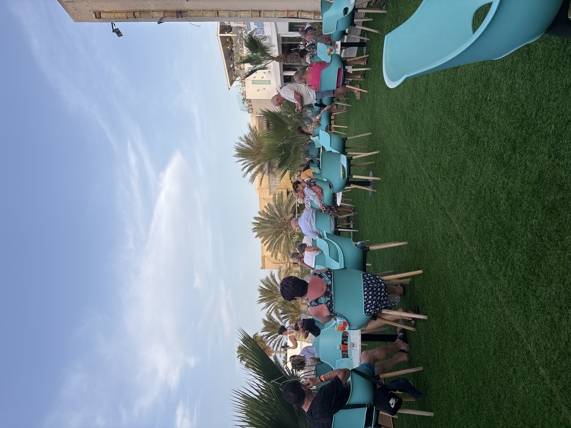 This image depicts an outdoor seating area with people dining and socializing. The area is covered with artificial grass and furnished with aqua-colored chairs around small, circular tables. In the background, buildings with light-colored facades and palm trees are visible under a partly cloudy blue sky. The scene appears to be a restaurant or cafe, likely in Taguermess, Tunisia, given the architecture and vegetation often found in coastal areas of Tunisia. Several individuals are seated at tables, some engaged in conversation, while others are holding drinks. One person stands near a table, holding a phone. The overall atmosphere suggests a relaxed, late afternoon or early evening gathering.