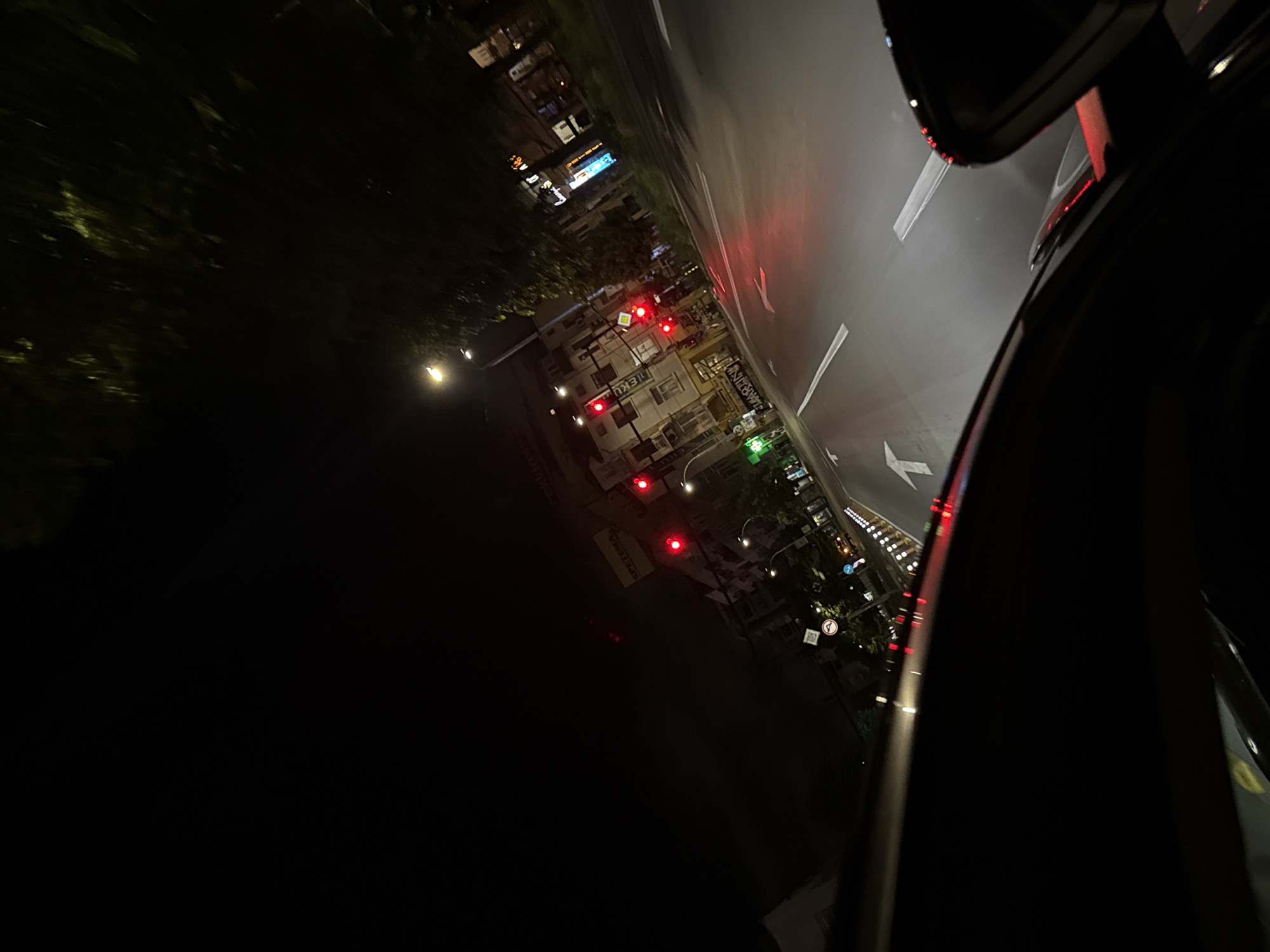 This image captures a nighttime street scene in Varna, Bulgaria, specifically at an intersection, viewed from inside a vehicle. The foreground reveals the dashboard and the reflection of a side mirror on the right. The vehicle's headlights illuminate the wet asphalt road ahead, which is marked with white arrows indicating straight or right-turn lanes. Multiple traffic lights are visible in the distance, all displaying red, indicating a stop.
Buildings line both sides of the street. On the left, a multi-story building features storefronts, including a pharmacy identified by a green cross sign and the text "APTEKA MARTIN" in Cyrillic. Further up on the same building, the sign "TEKU" is visible. On the right side of the street, another building prominently displays a "SPETEMA" sign, indicating a coffee shop or similar establishment. Overhead streetlights cast bright illumination onto the scene, reflecting on the damp road surface. Trees with dark foliage are visible along the right side of the street, partially lit from below by streetlights. No individuals are distinctly visible, and no active interactions or actions are depicted beyond the static nighttime urban environment.