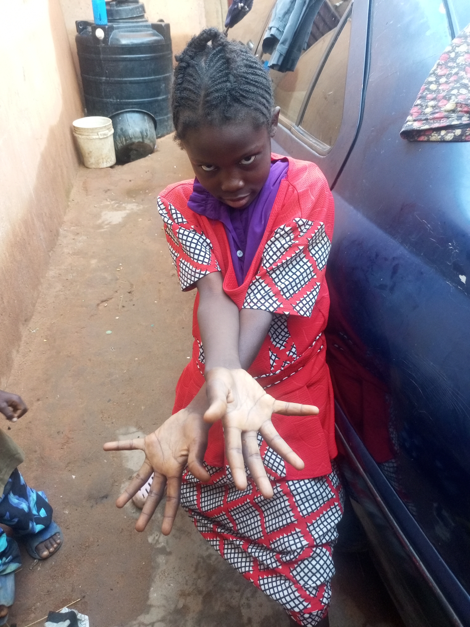 A young Black girl with braided hair, wearing a purple top and a red and white patterned dress, stands in front of a dark blue vehicle. She is extending her hands towards the camera, palms open. In the background, a large black water tank and a white bucket are visible against a sandy-colored wall. The scene appears to be an outdoor space, possibly a courtyard or alleyway, in Tempure, Nigeria. The ground is dirt and there is some debris present.