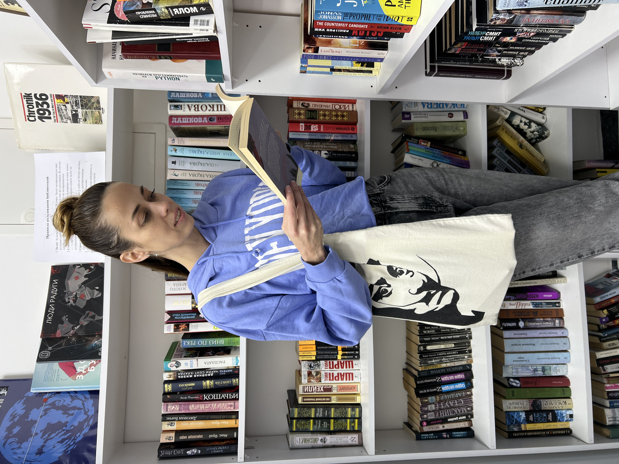 A woman with a light blue hooded sweatshirt, gray jeans, and a white tote bag is sitting or lying in a white bookcase. The bookcase is full of books. She is holding a book and is looking at it. The bookcase is in an indoor setting. The location is Bar, Montenegro.