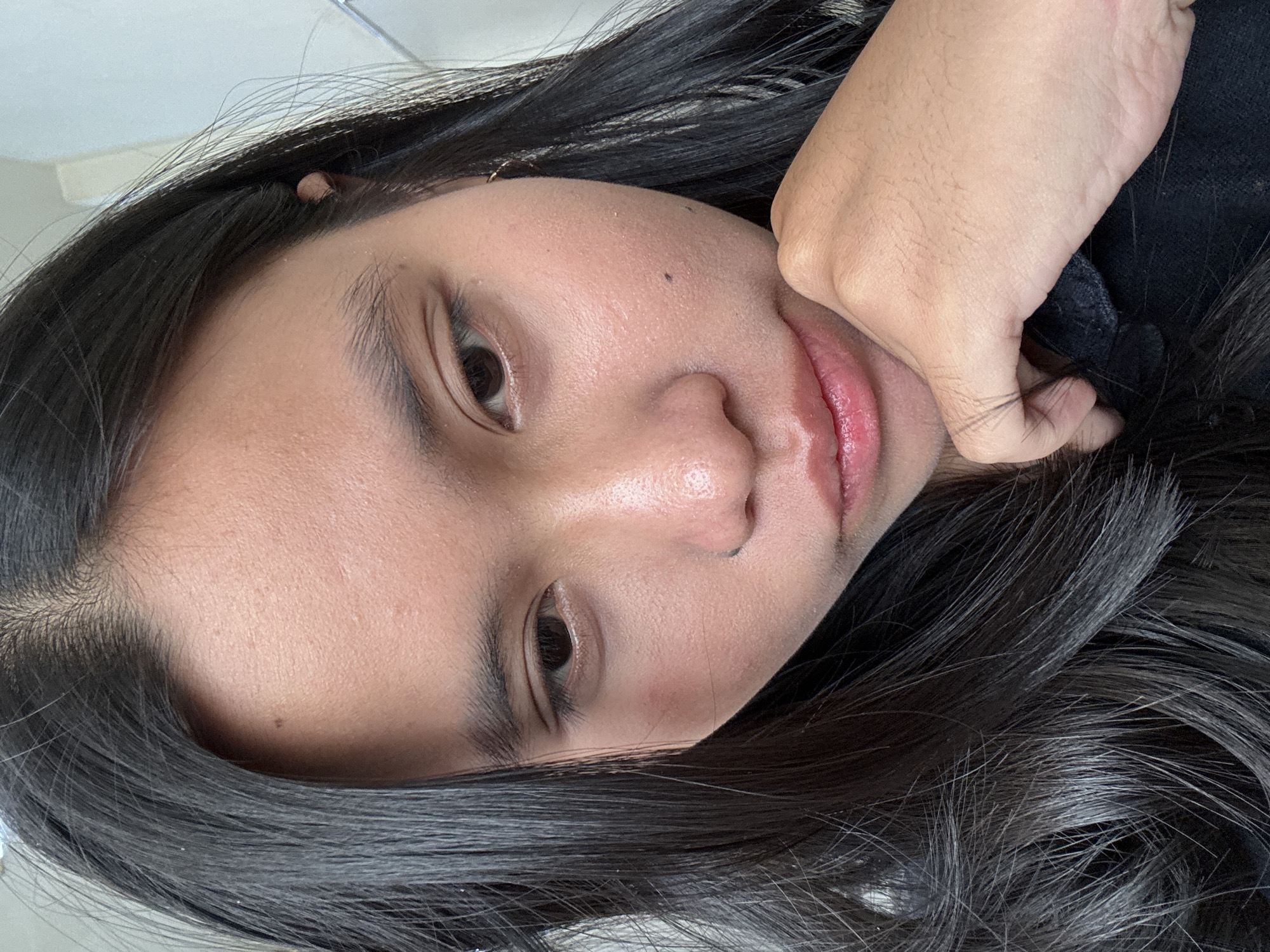A medium-close-up, vertically oriented selfie shows the face of a young person of Southeast Asian descent. The person's head is tilted down and slightly to the left, with their chin resting on their left hand. Dark, long hair frames the face and drapes over the shoulder. The skin appears to have a light sheen, and there are a few small blemishes. The person's eyes are looking downwards. Their lips are slightly parted and appear to have a pink tint. The background is out of focus, with a hint of a light-colored wall and possibly some vertical lines suggesting a window or blinds. The image appears to have been taken indoors, in what could be a home or office environment. It is not possible to determine the specific location within East Jakarta, Indonesia, from the visual information provided in the image.