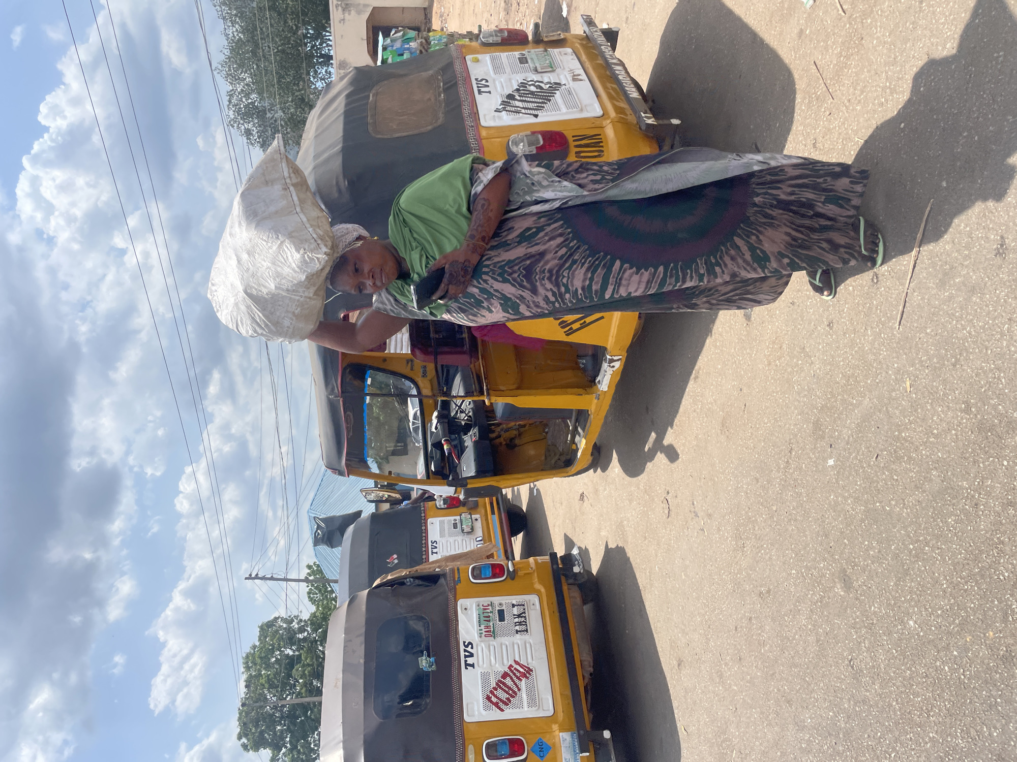 A woman with dark skin carries a large white sack on her head as she stands next to a yellow motorized tricycle (auto-rickshaw) on a paved street. She wears a light green shirt, a patterned dark and light brown or gray wrap skirt, and sandals. Her arm is decorated with henna-like patterns. She holds a small black object in her hand, possibly a phone. The tricycle is parked in a line with other similar tricycles. The license plate on the tricycles is partially visible, featuring the text "TVS" and other details. The background shows utility poles with wires, a building, and a cloudy sky. Based on the vehicles and the style of dress, it is highly probable that the image was taken in Lokoja, Nigeria.