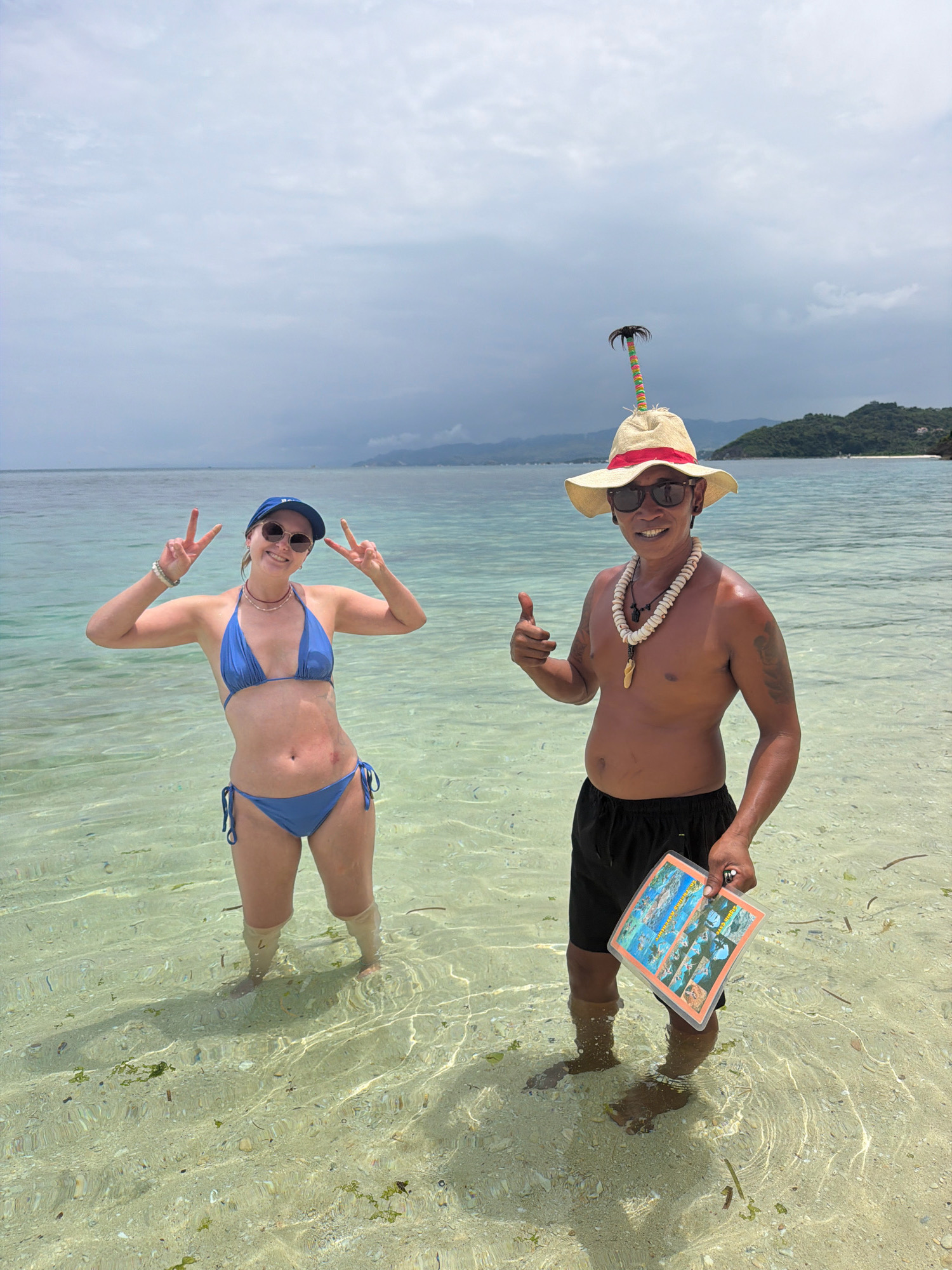 A woman in a blue bikini and a man in black shorts and a straw hat with a palm tree decoration are standing in shallow, clear turquoise water on a beach in Malay, Philippines. The woman is smiling, wearing a blue baseball cap and sunglasses, and making a peace sign with both hands. The man is also smiling, giving a thumbs-up, and wearing a shell necklace. He is holding a plastic-covered pamphlet with pictures. In the background, the sea stretches to the horizon with a distant, green, tree-covered island. The sky is overcast. The seabed is visible through the clear water, with sand and some seaweed.