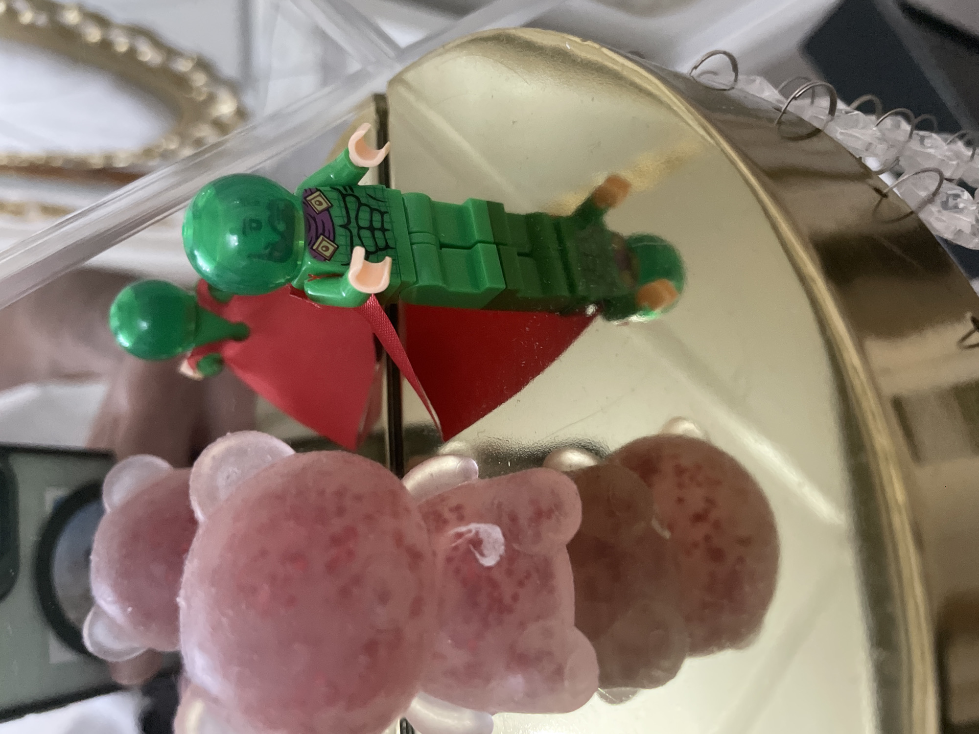 An angled, close-up shot captures a collection of items on and reflected by a golden, circular surface. The main subjects are a Lego minifigure and several bear-shaped objects.
The Lego minifigure, positioned at the top, features a vibrant green head with etched facial details, a green body with purple design elements on its torso, and a striking red cape. Its clear reflection is visible on the highly polished golden surface directly beneath it.
Below the minifigure, a row of translucent, pinkish-red bear-shaped objects is arranged. These objects, possibly candies, have small, darker specks on their surface. Their reflections are also clearly visible on the reflective golden base.
In the softly blurred background, a golden, ornate object with a decorative, possibly scalloped, edge is partially visible. To the right, a clear plastic structure with a series of small, silvery rings is present, suggesting a display or storage component. The setting appears to be indoors, likely on a shelf or within a display case. No text is visible in the image. The scene is well-lit, highlighting the reflective properties of the golden surface and the textures of the objects. The location is Ihtiman, Bulgaria.
