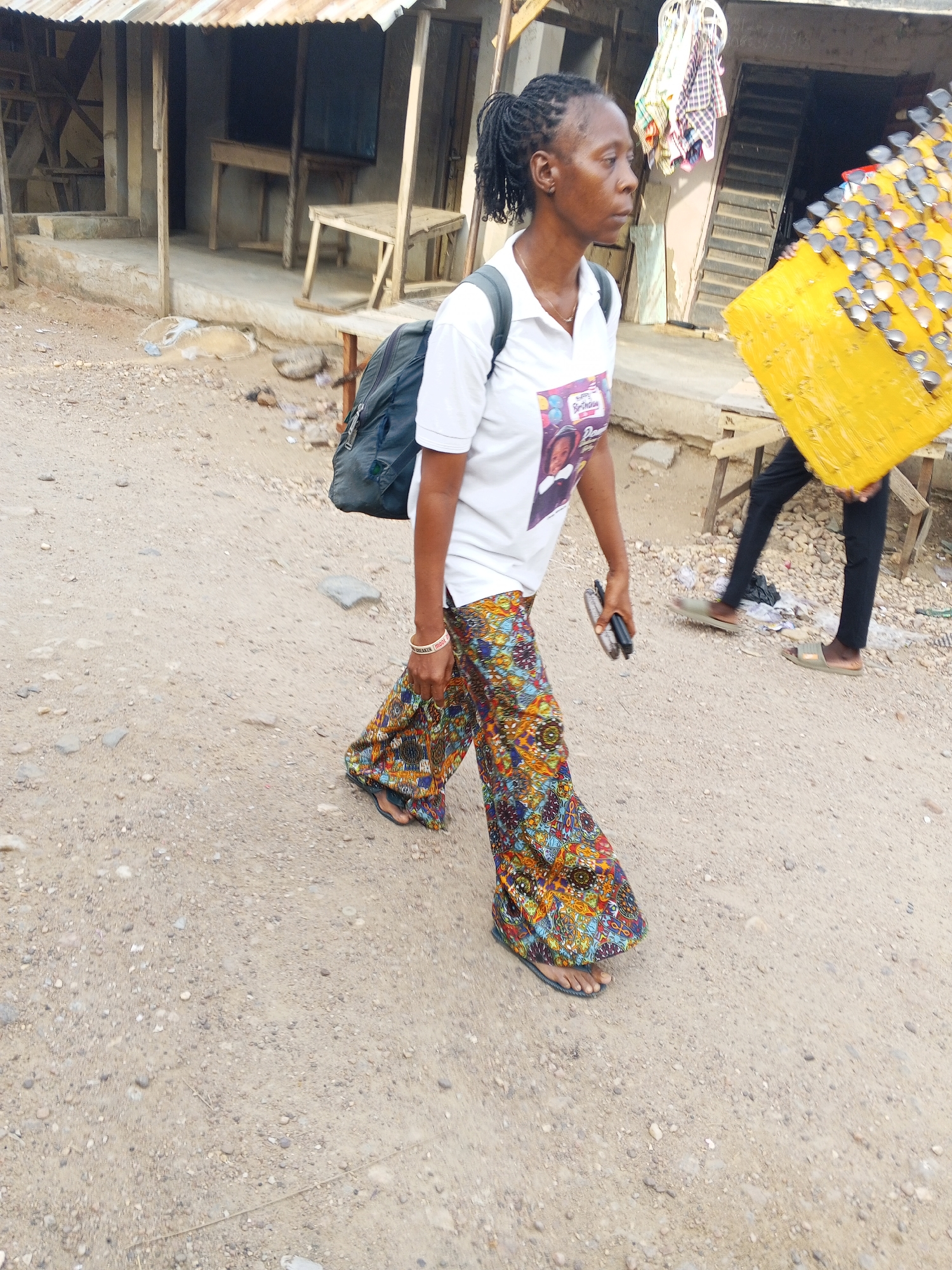 A woman with braided hair, wearing a white t-shirt, colorful patterned pants, and flip-flops, walks on a dirt road in Adankolo, Nigeria. She carries a black backpack and something in her hand. A person walks beside her carrying a yellow rectangular object covered in sunglasses. Dilapidated wooden structures and benches are visible in the background. The ground is dirt and gravel.