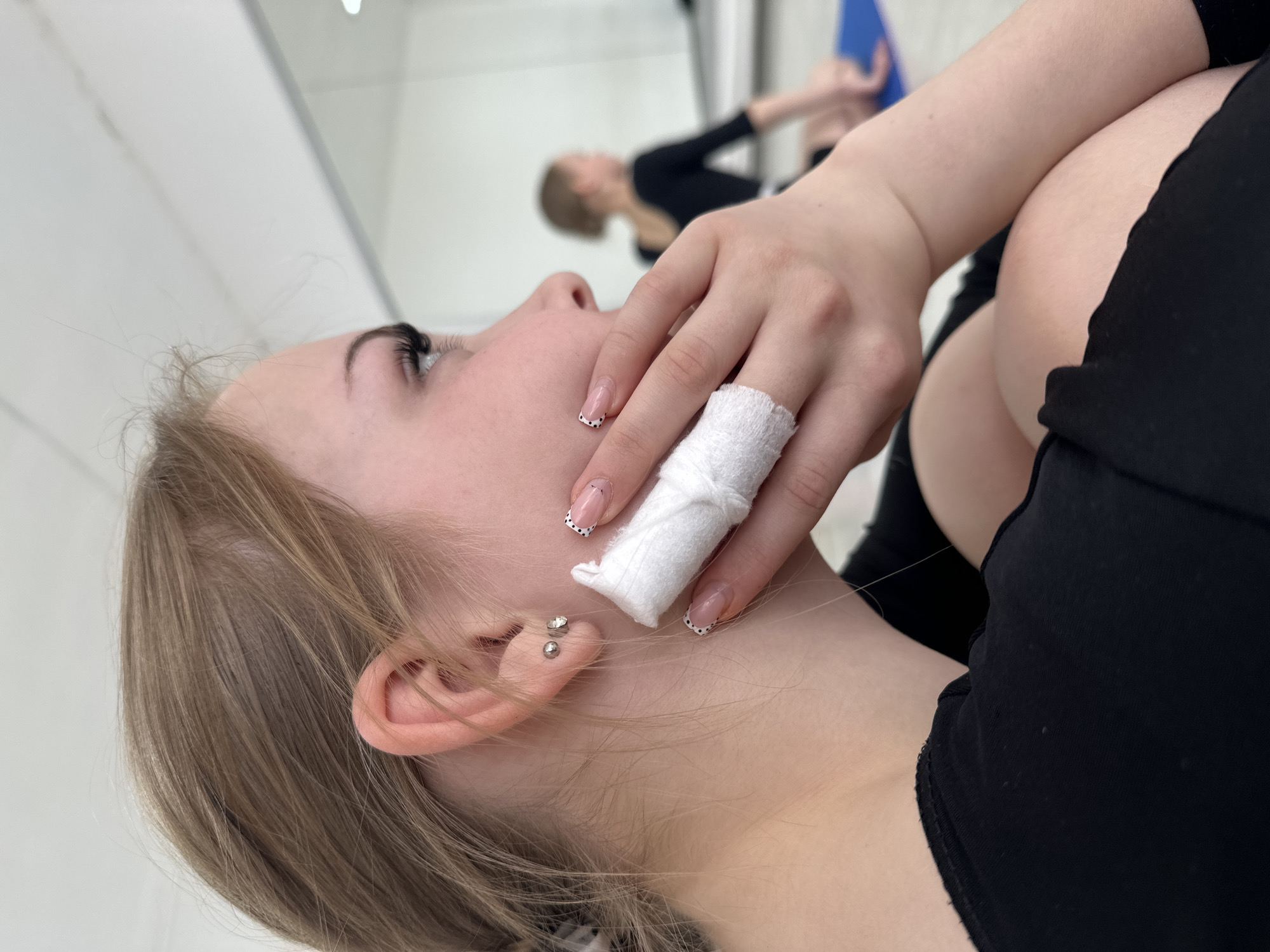 A close-up, high-angle shot captures a young woman in the foreground, lying or reclining with her head tilted back, looking upwards. She has fair skin, light brown hair, and long eyelashes. Two small, silver ear piercings adorn her earlobe and helix.
Her left hand is prominent in the frame, held near her face. One of her fingers, likely the index finger, is wrapped in a white bandage or cloth, possibly indicating a minor injury or a specific activity requiring a finger protector. Her fingernails are manicured with a French tip style, featuring small black dots along the white tips. She is wearing black clothing, appearing to be a top or leotard, and possibly leggings or trousers, as suggested by the fabric near her knee.
In the blurred background, a reflection in a large mirror shows another individual, who appears to be the same young woman, standing or moving in a bright, white-walled room. This reflected figure is also wearing black attire and holds a blue object, possibly a folder or mat. The setting suggests an indoor studio, likely for dance or fitness activities, given the mirror and attire. The overall mood is calm, with the foreground subject seemingly at rest. There is no visible text in the image.